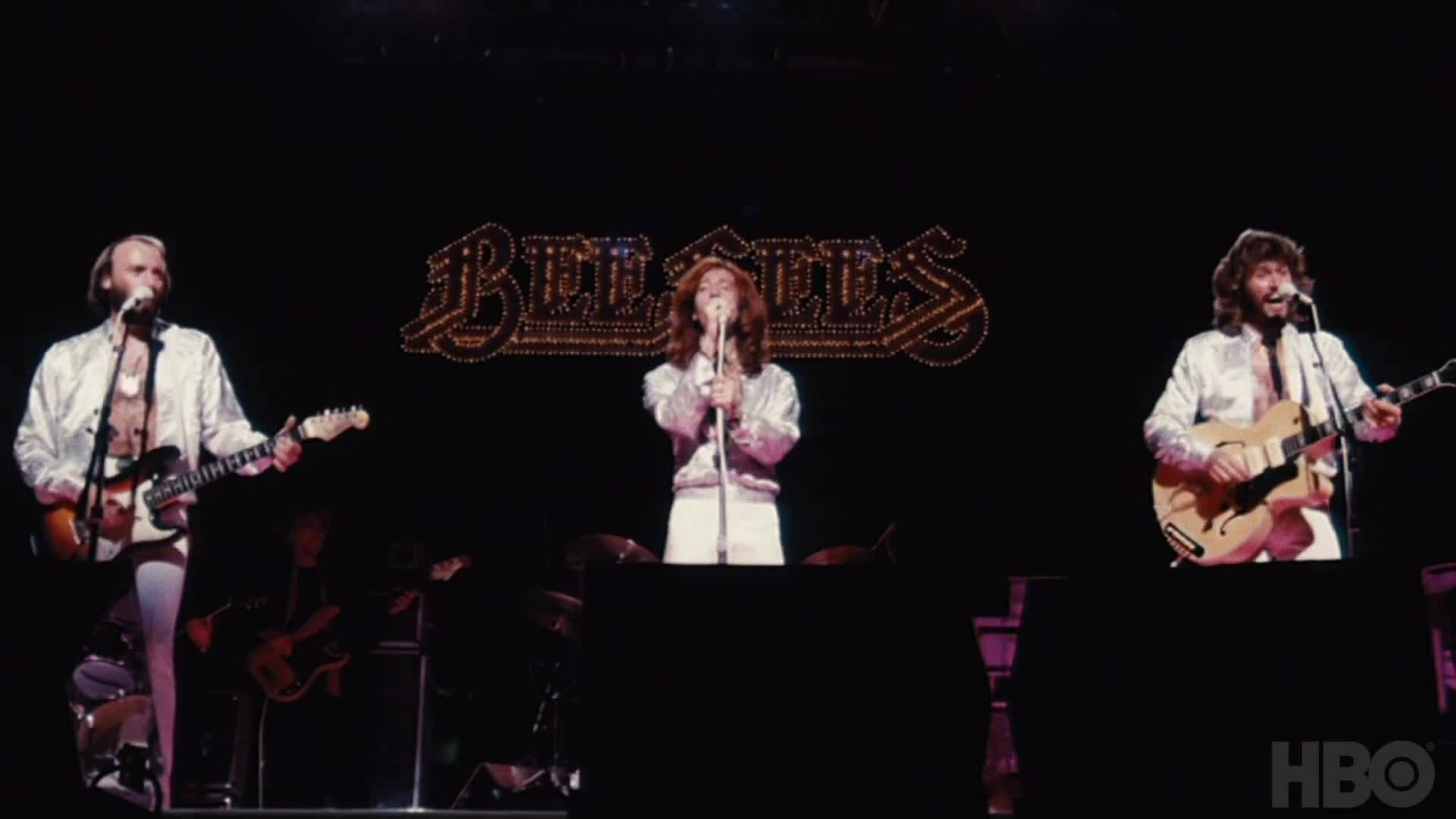 Celebrity 桌布 壁紙 — 三位身穿白色服裝的音樂家在閃閃發光的 Bee Gees 標誌下，手持吉他和麥克風在舞台上表演。