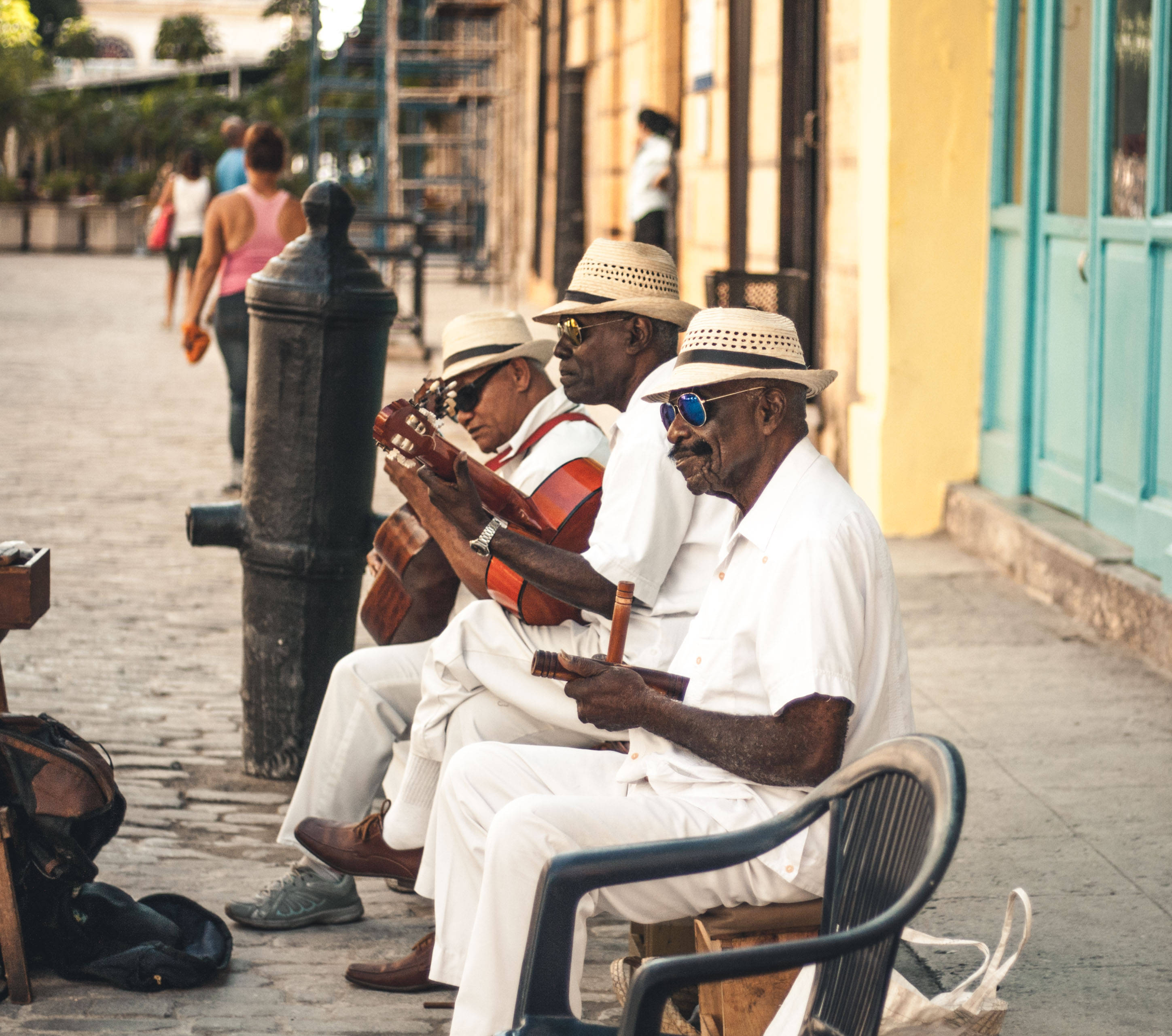 Cuba 桌布 壁紙 — 三位身穿白色西裝、戴草帽的年長古巴音樂家在陽光明媚的鵝卵石街道上彈吉他。
