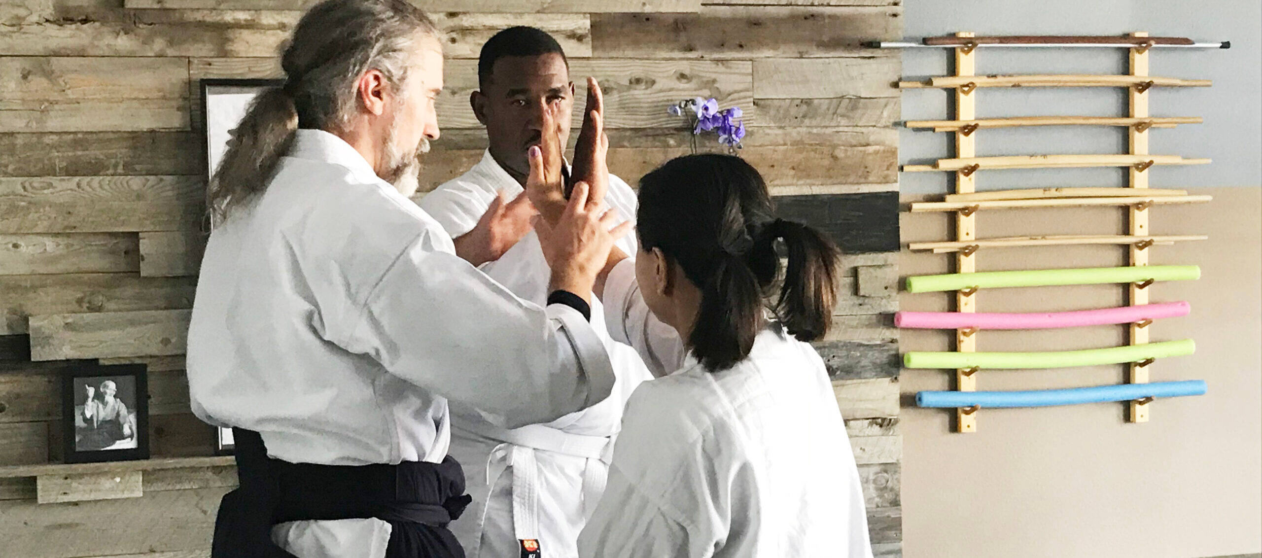martial arts 桌布 壁紙 — 三位身穿白色道服的武術家，在質樸的木牆前，用色彩繽紛的訓練棍練習精準的手部技巧。