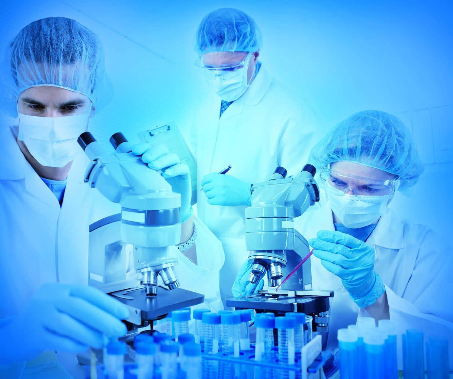 科學 桌布 壁紙 — 三位身穿白袍和口罩的科學家在無菌的藍色燈光實驗室中，專注地用顯微鏡檢查樣本。