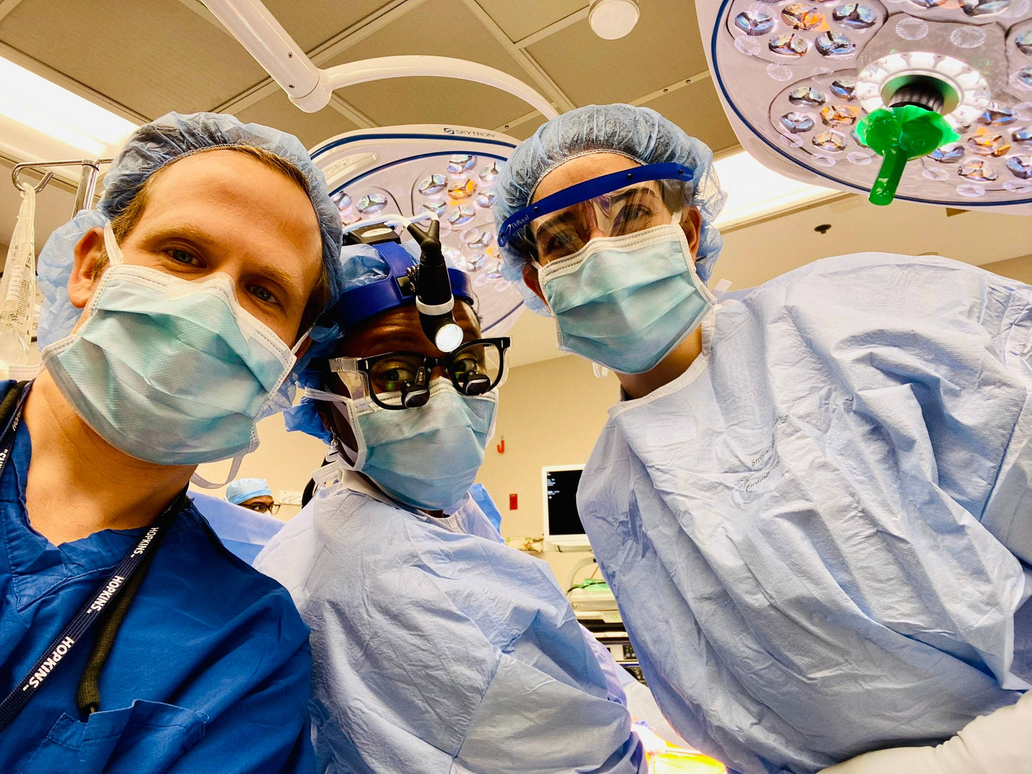 Medical 桌布 壁紙 — 三位身穿藍色手術服、口罩和帽子的外科醫生在明亮手術燈下於手術室中對著鏡頭微笑。