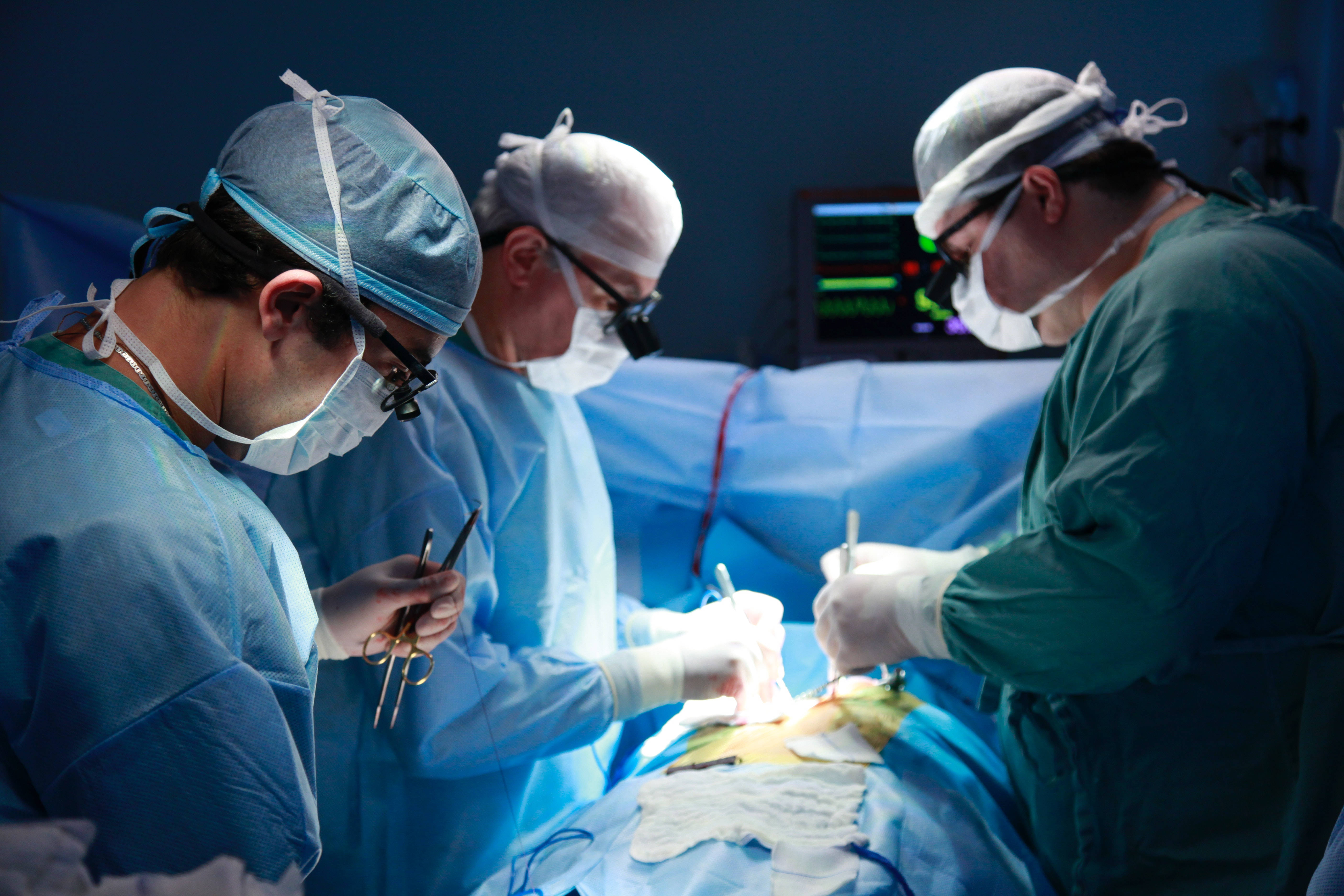 Surgeon 桌布 壁紙 — 三位身穿藍色手術服和口罩的外科醫生，在無菌手術室明亮的手術燈下進行精細的手術。