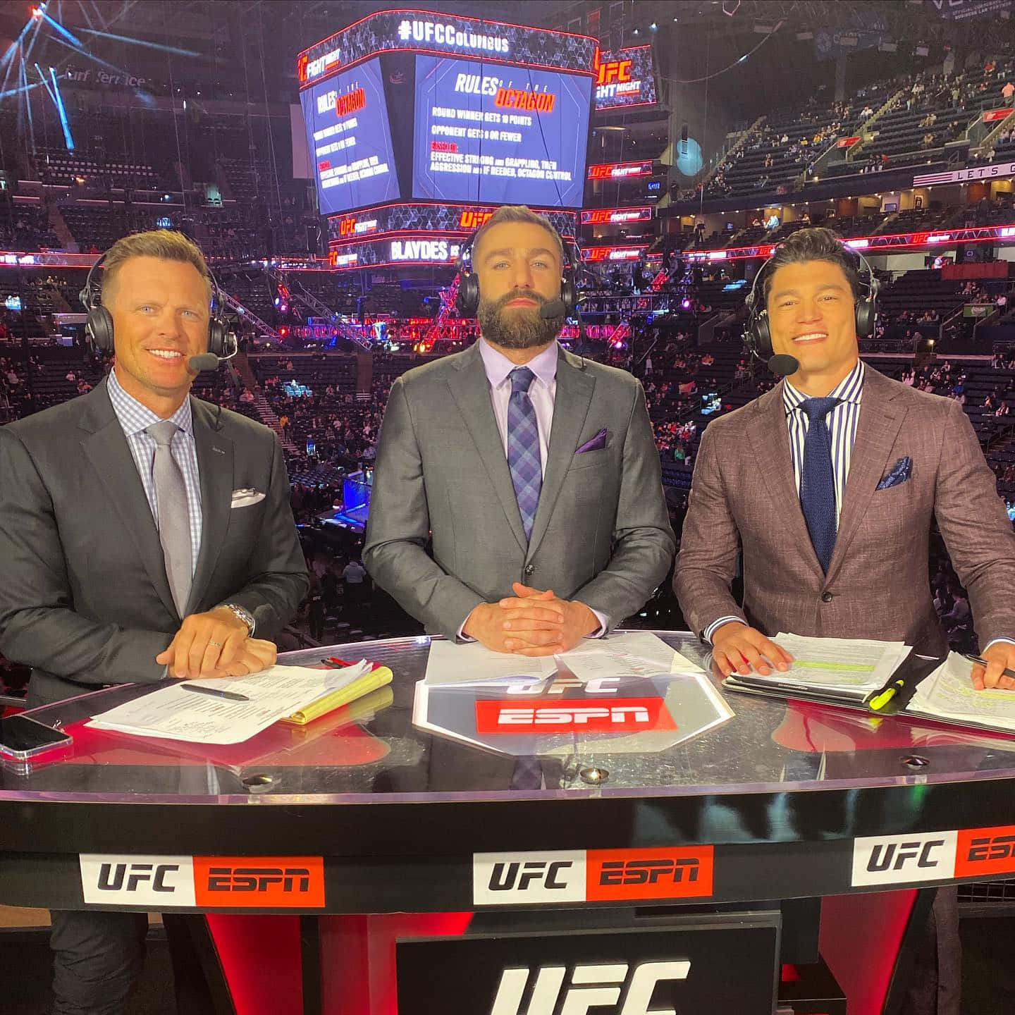 UFC 桌布 壁紙 — 三位身穿西裝、面帶微笑的男性評論員坐在UFC ESPN的轉播桌前，身後是座無虛席的競技場和發光的螢幕。