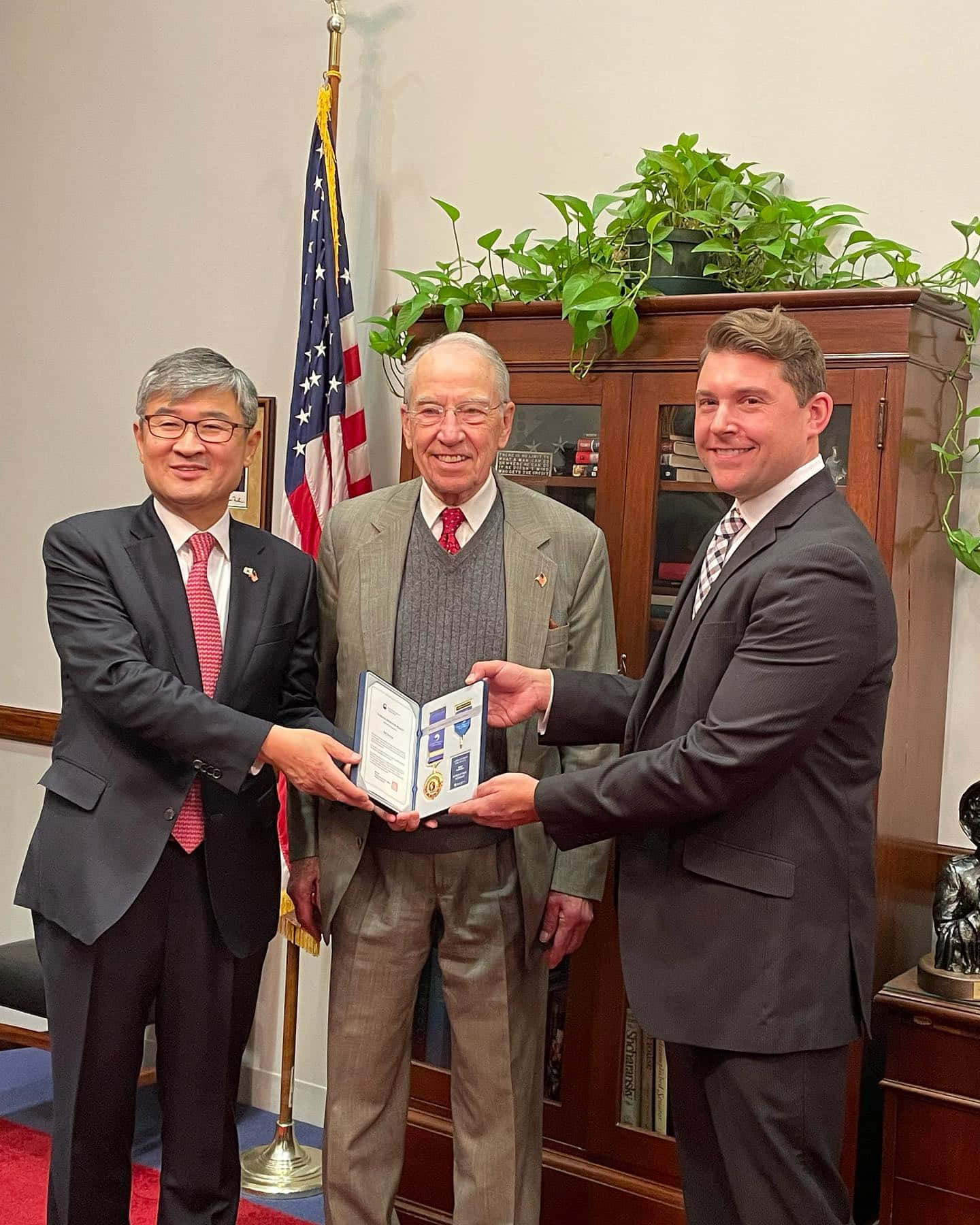 政治 桌布 壁紙 — 三位身穿西裝的男士在一個正式辦公室內交換一個裱框證書，辦公室內有美國國旗和木製家具。