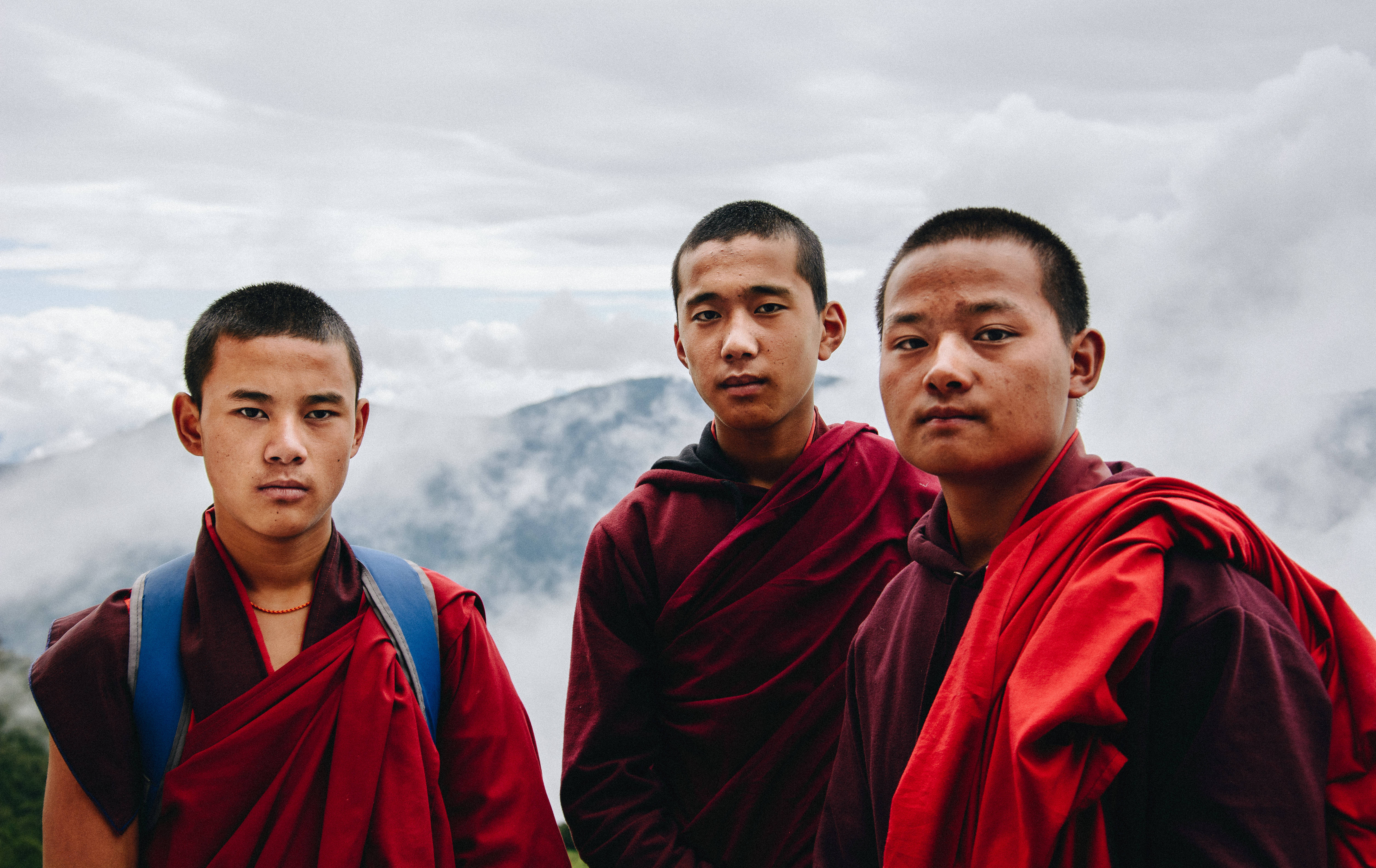 Bhutan 桌布 壁紙 — 三位身穿鮮紅色僧袍的年輕佛教僧侶，在陰天之下，以霧氣繚繞的山巒為背景，莊嚴地站立著。