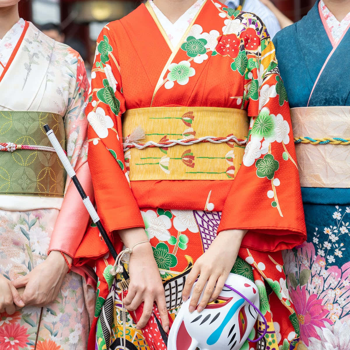 Kimono 桌布 壁紙 — 三位身穿鮮豔傳統和服、帶有花卉圖案的女士站在一起，手持裝飾面具。