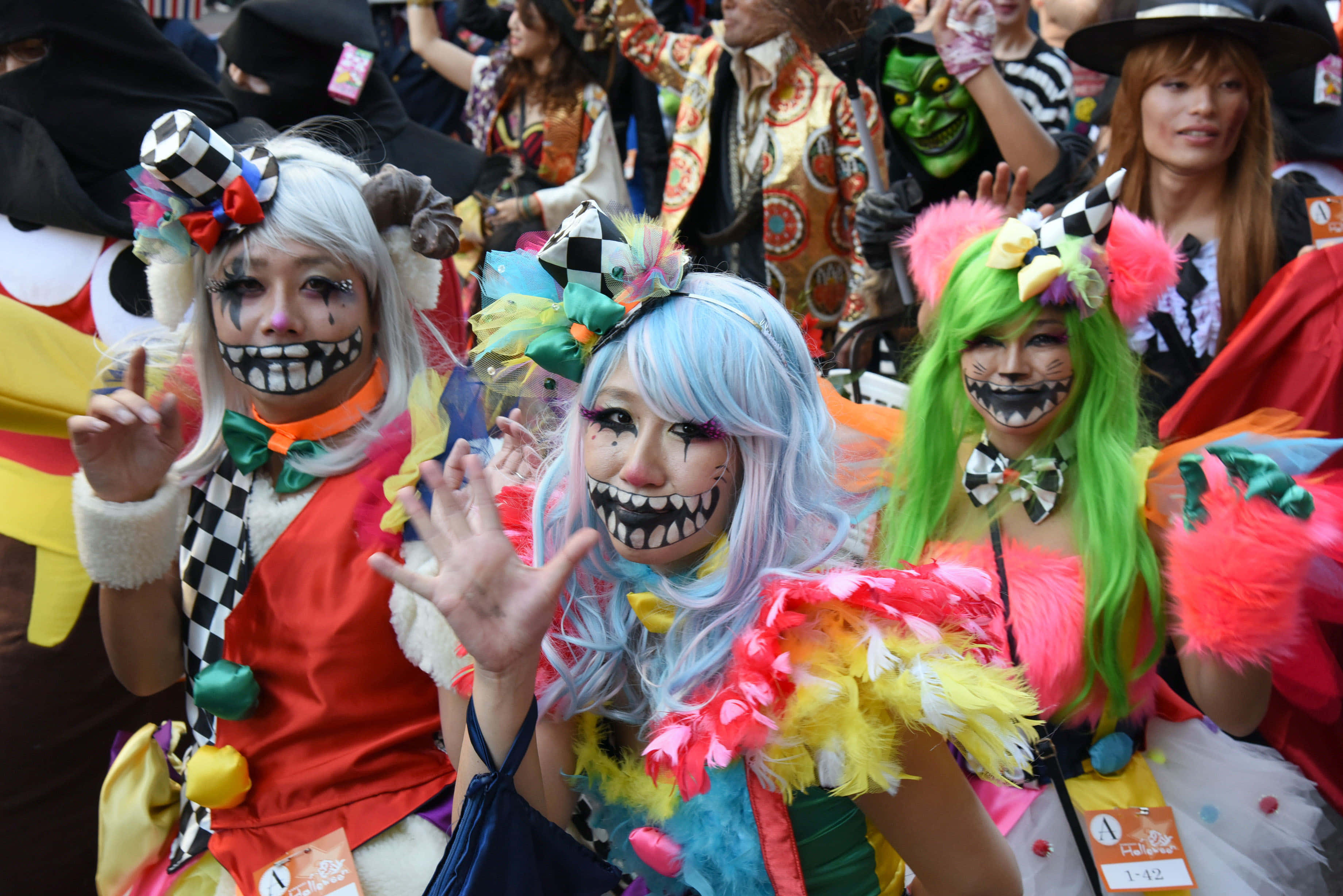 Halloween Parades 桌布 壁紙 — 三位身穿鮮豔小丑服裝、塗著牙齒、戴著彩色假髮的女性在熱鬧的節日中向鏡頭揮手。