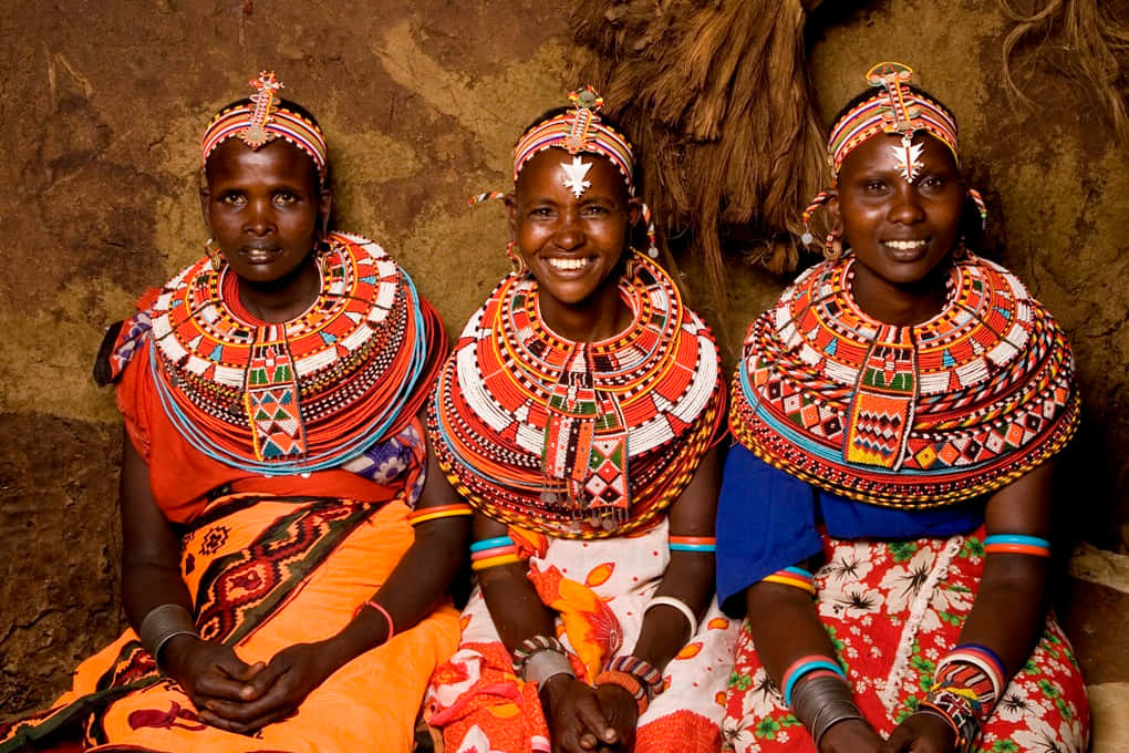 African Culture 桌布 壁紙 — 三位身穿鮮豔珠飾服裝的馬賽婦女，在質樸的土色牆壁前溫暖地微笑。