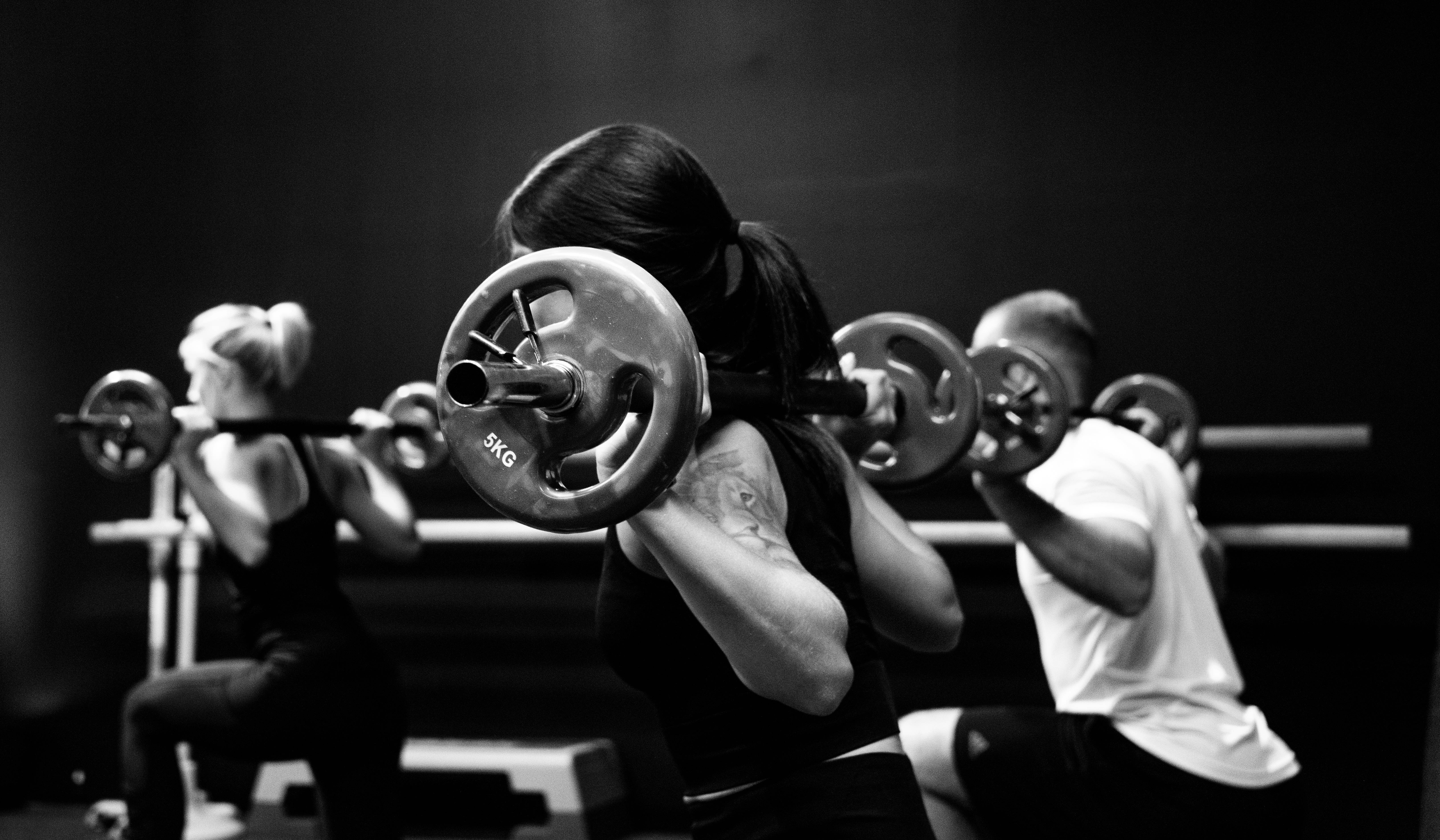 Fitness 桌布 壁紙 — 三位運動員在昏暗的健身房中舉起沉重的槓鈴，呈現強烈的黑白對比效果。