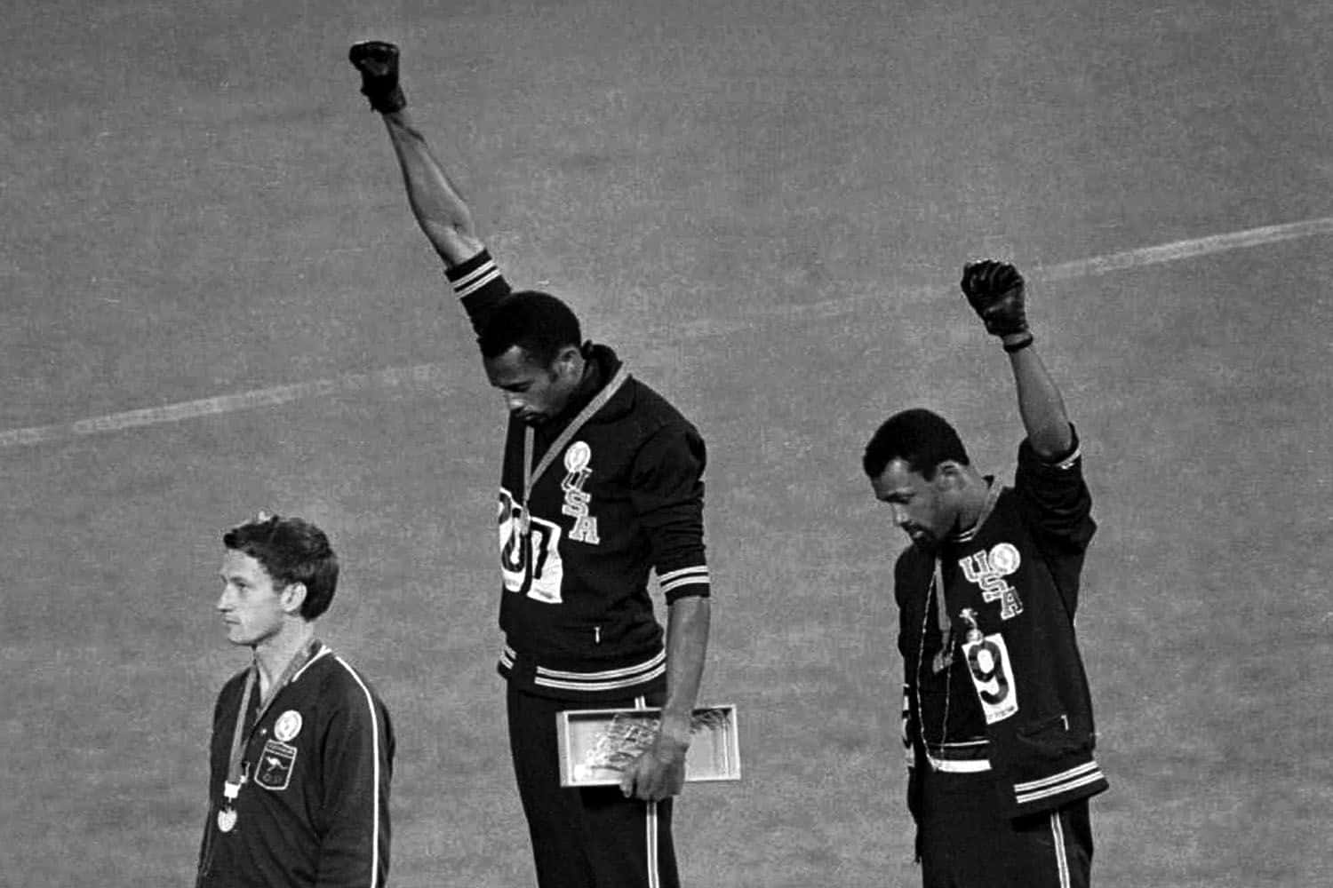 Black Power 桌布 壁紙 — 三位運動員站在頒獎台上，其中一人舉起拳頭抗議，另一人則莊嚴地注視著，這是一張歷史性的黑白照片。
