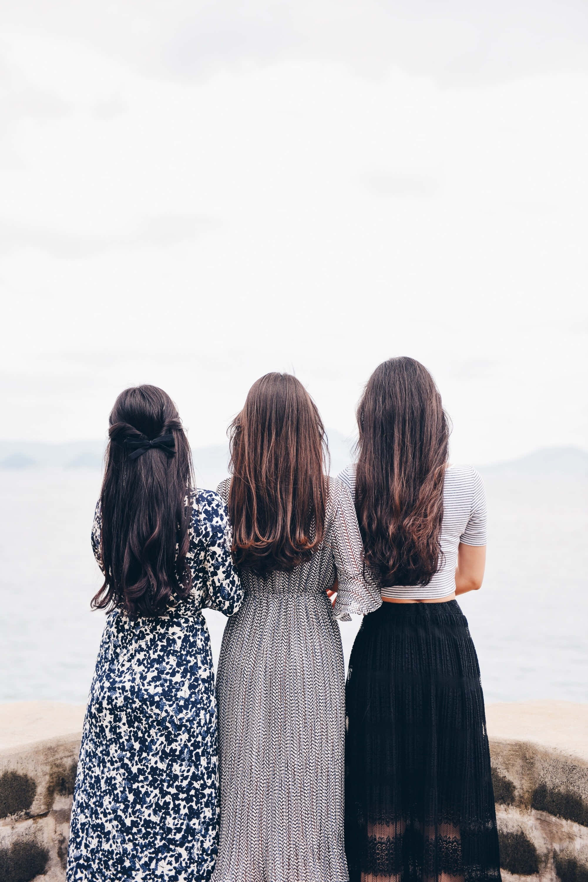 women 桌布 壁紙 — 三位長髮女性背對背站在岩石海岸上，凝視著多雲天空下平靜的大海。