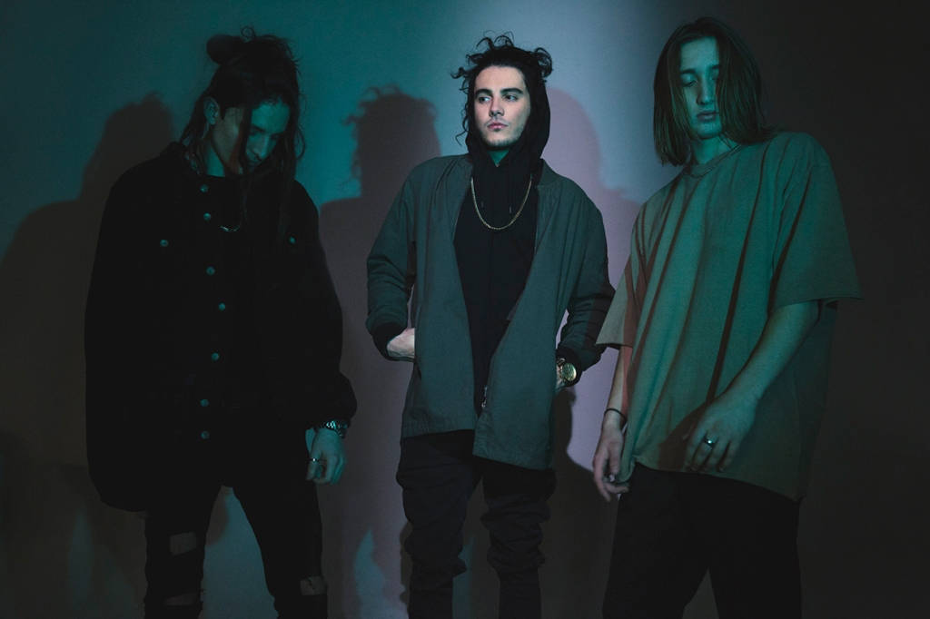 Chase Atlantic 桌布 壁紙 — 三位長髮年輕男子站在昏暗的房間中，沐浴在青綠色和紫色的燈光下，在他們身後的牆上投下戲劇性的陰影。