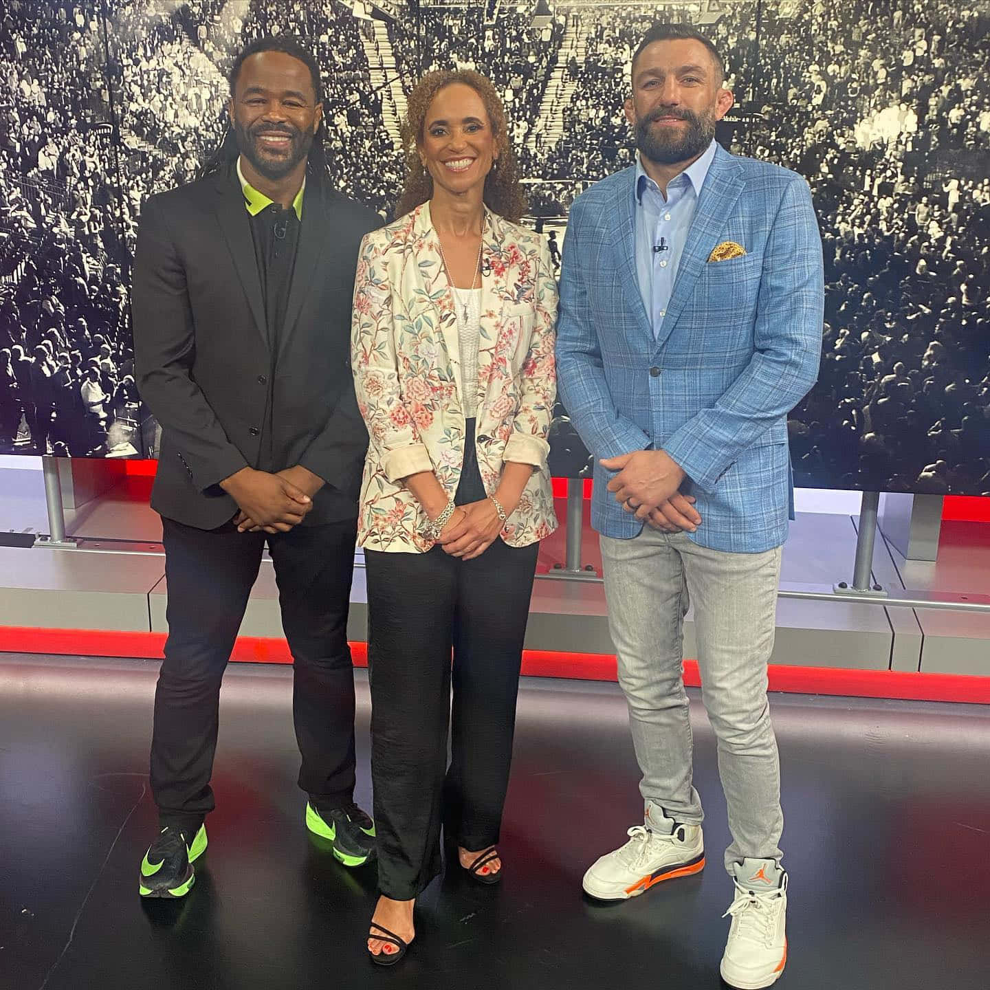 運動 桌布 壁紙 — 三位面帶微笑的主持人身著時尚服裝，在現代攝影棚的黑白人群背景前自信地擺姿勢。