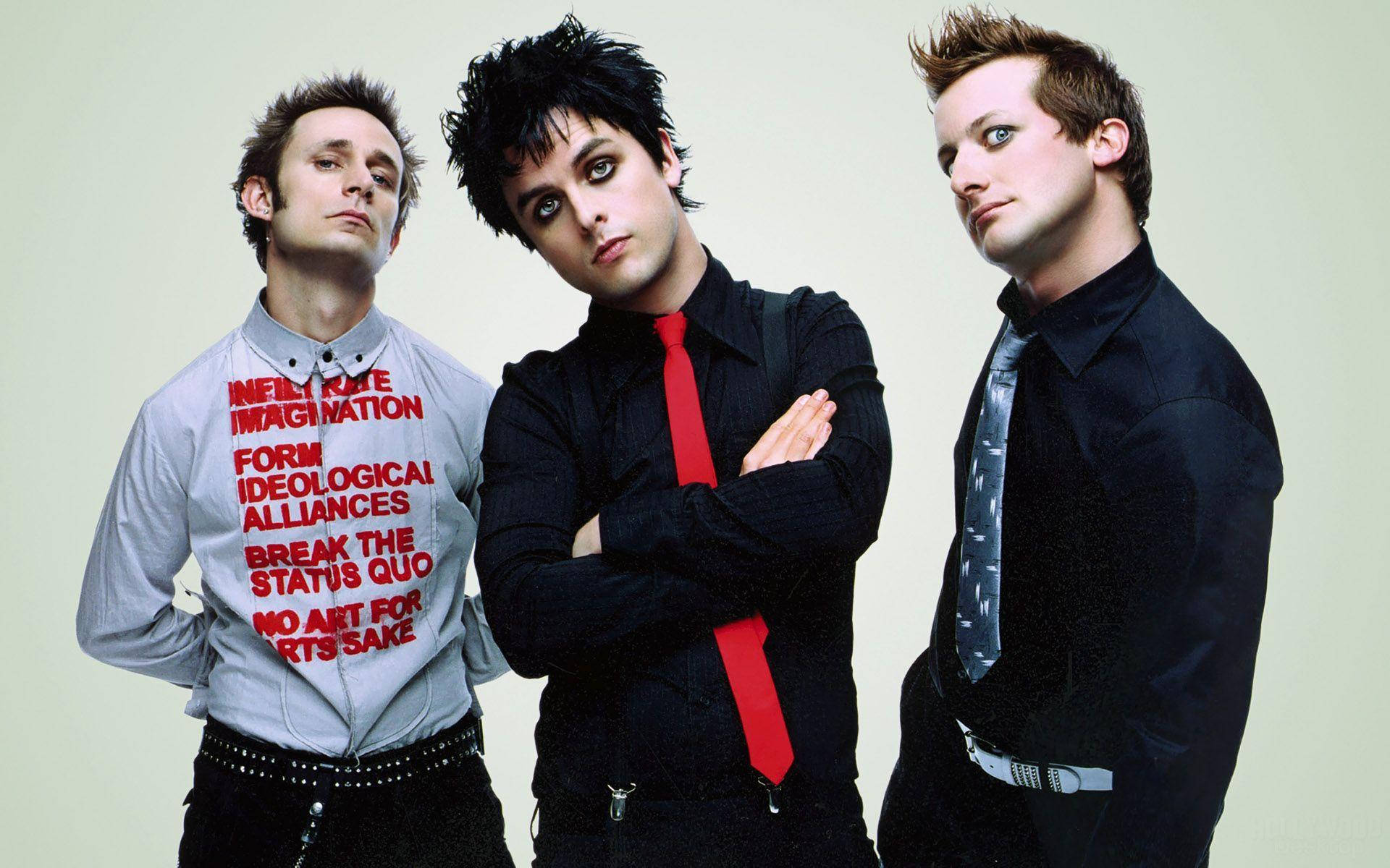 Green Day 桌布 壁紙 — 三位龐克搖滾音樂家在素色背景前自信地擺姿勢，其中一人穿著印有大膽紅色政治標語的襯衫。