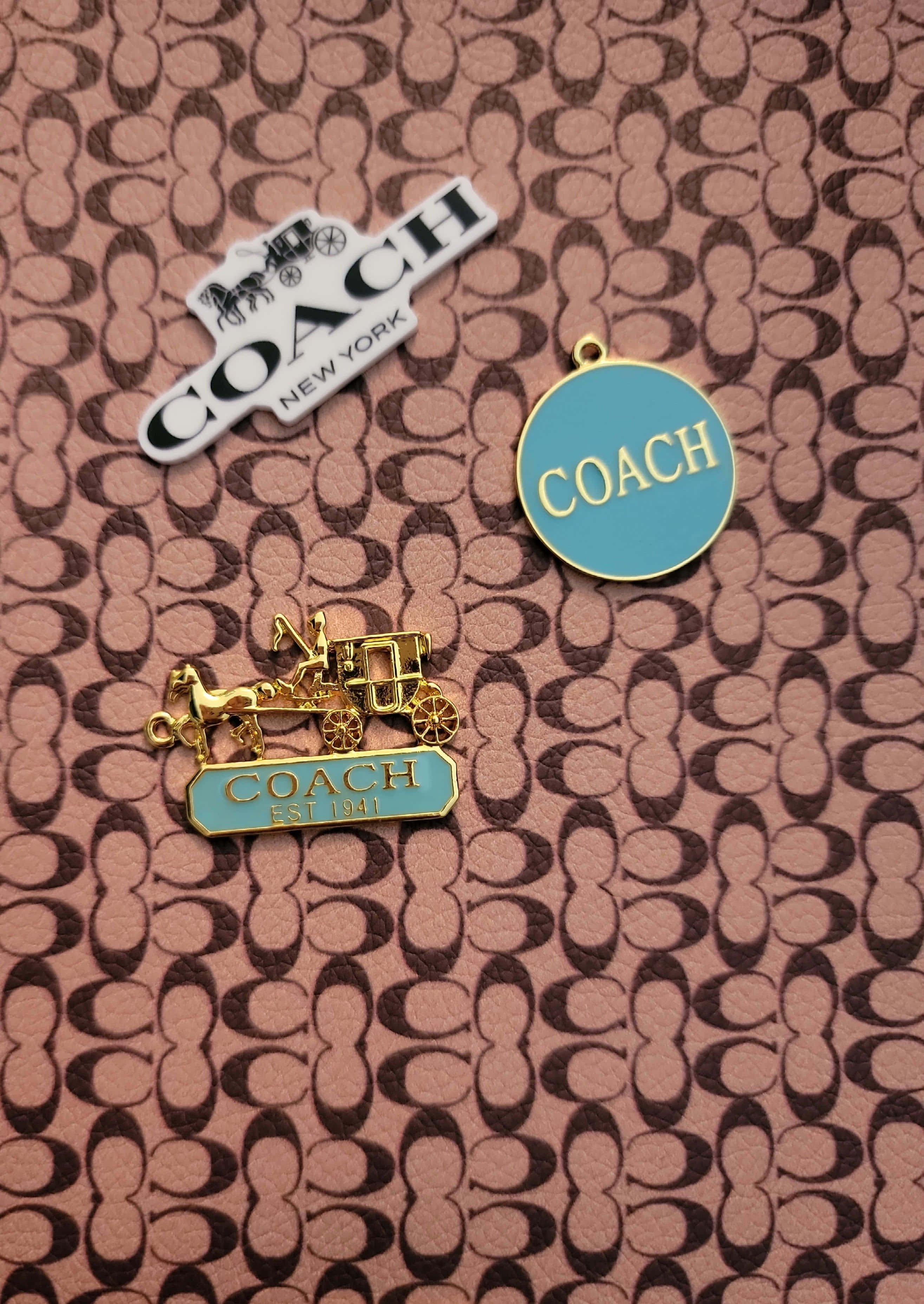 Coach 桌布 壁紙 — 三個Coach品牌別針放置在帶有紋理的粉紅色和棕色標誌性圖案皮革表面上。