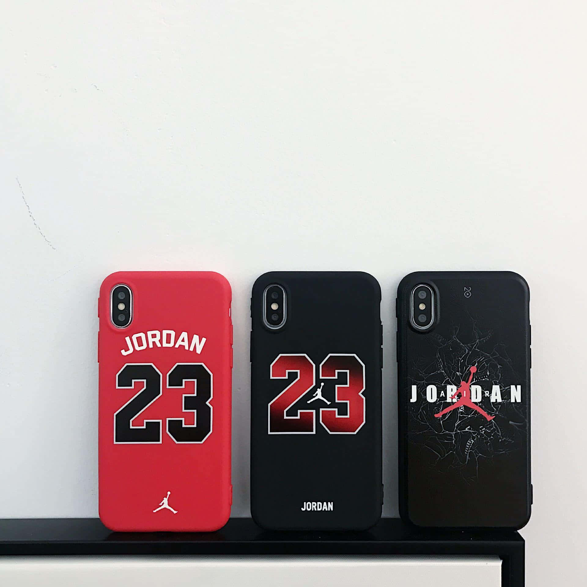  壁紙 — 三個Jordan 23手機殼，分別為紅色、黑色和裂紋黑色設計，整齊地放在一個白色牆壁前的架子上。