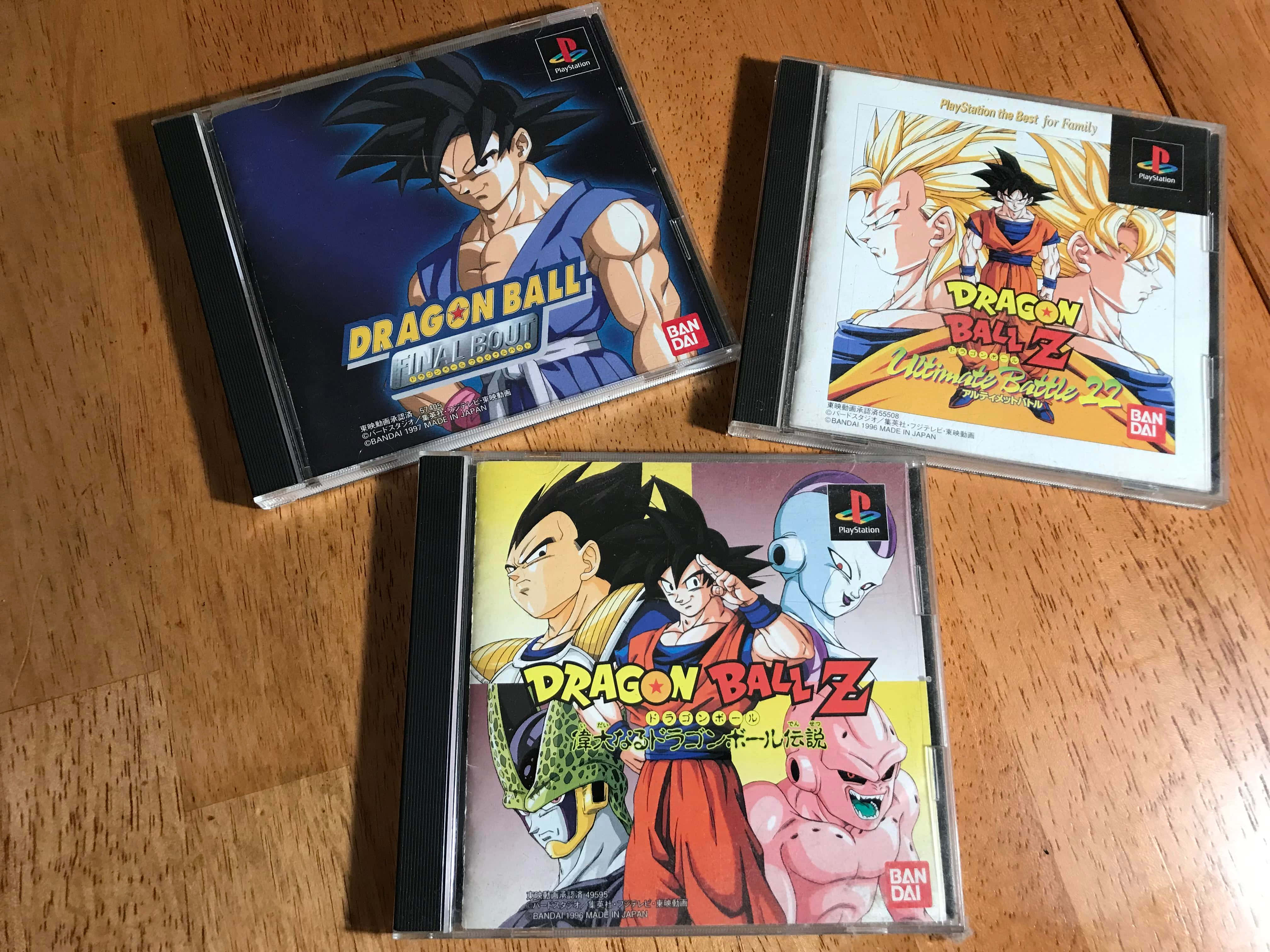 Dbz Video Games 桌布 壁紙 — 三個七龍珠Z PlayStation遊戲盒整齊排列在溫暖的木地板上，展示充滿活力的動漫角色封面藝術。