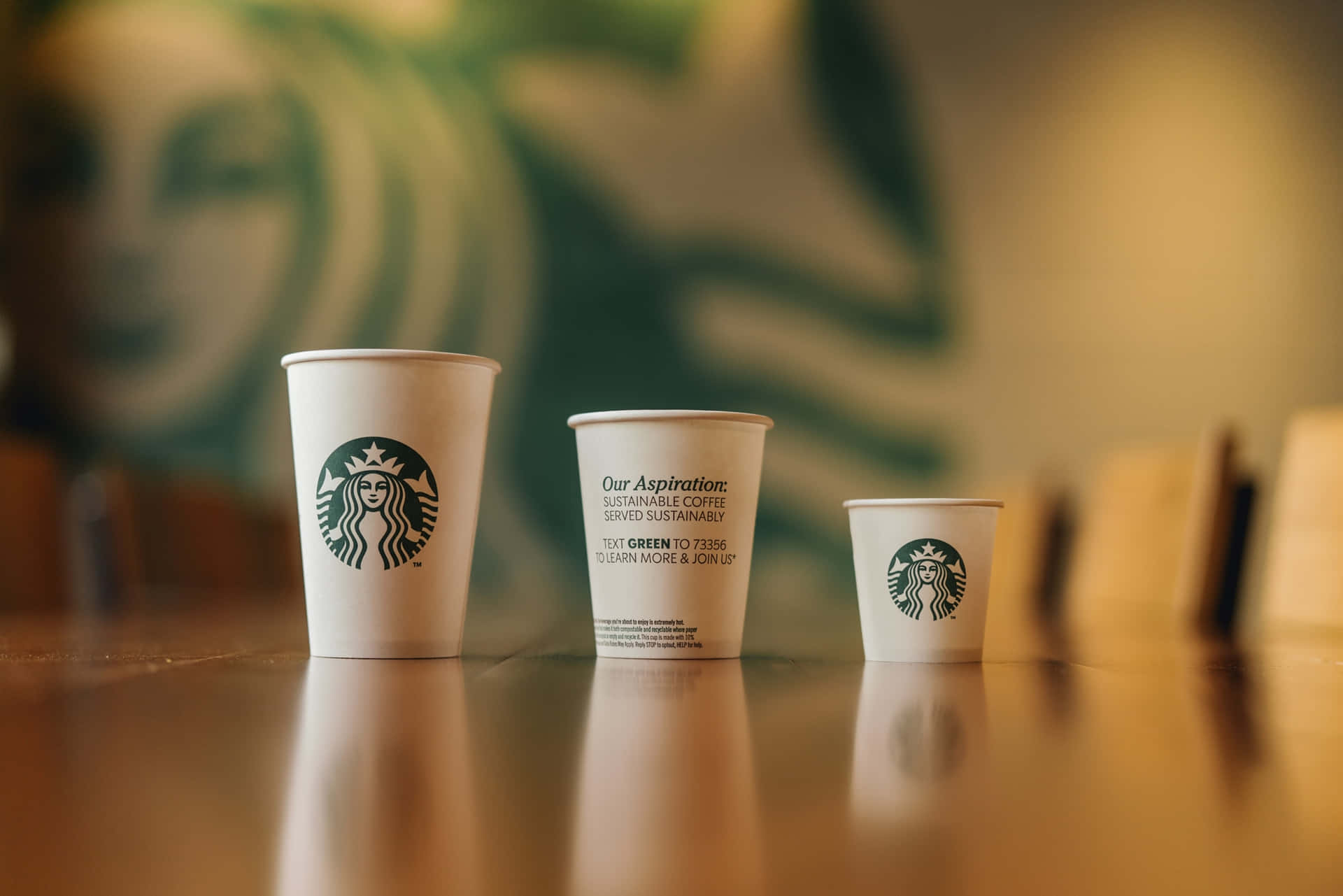 星巴克 桌布 壁紙 — 三個不同大小的Starbucks咖啡杯放在木製櫃檯上，背後有模糊的綠色標誌牆。