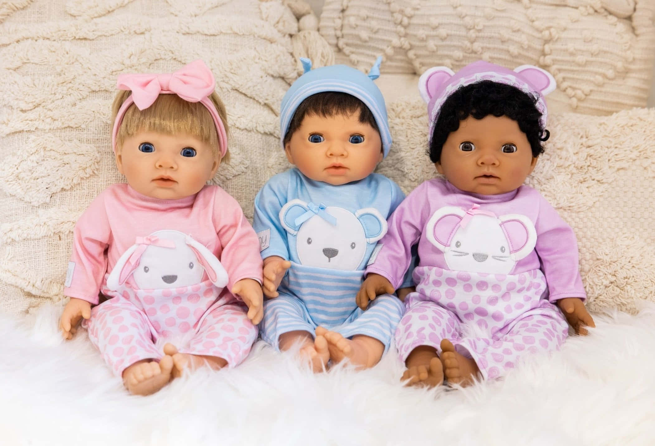 Doll 桌布 壁紙 — 三個不同的嬰兒娃娃穿著粉彩服裝，並排坐在毛絨白色地毯上，背景是帶有紋理的奶油色背景。