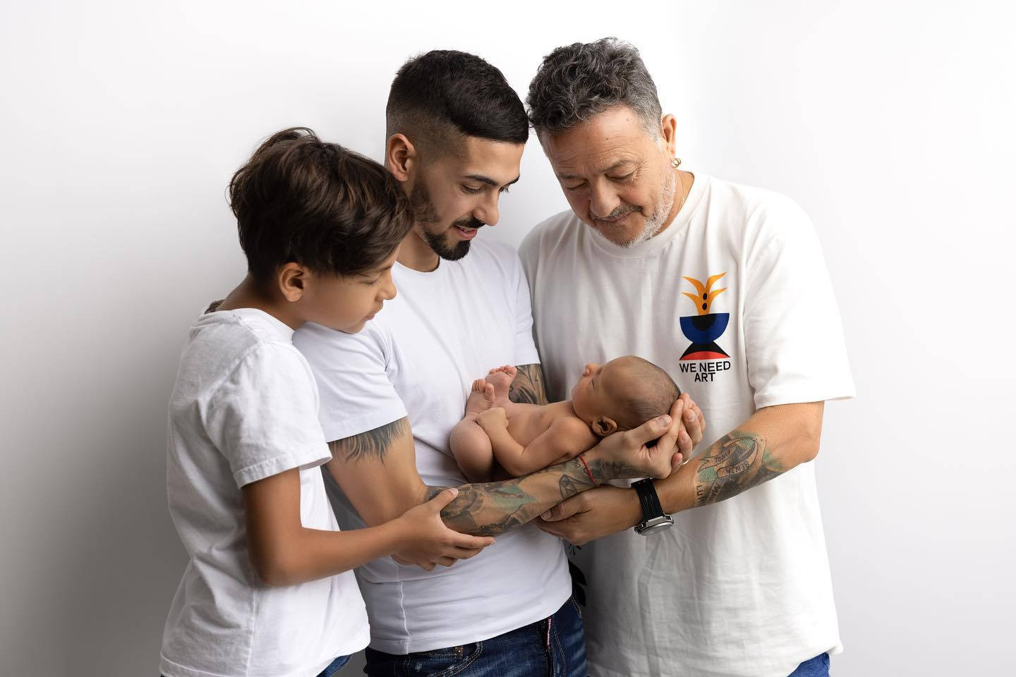 Family 桌布 壁紙 — 三個世代的男人穿著白襯衫，在純白背景前輕輕懷抱著一個熟睡的新生兒。