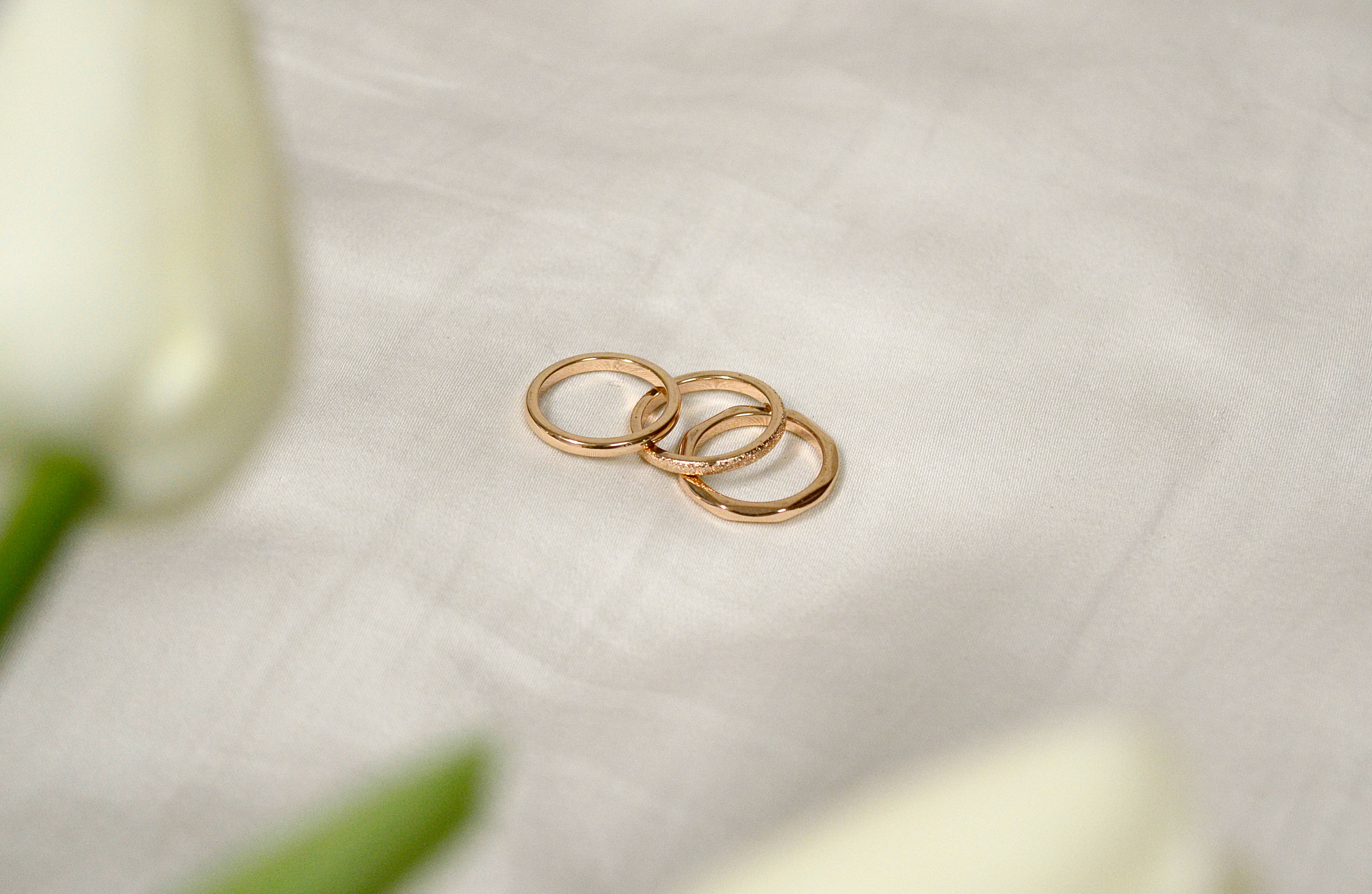 玫瑰金 桌布 壁紙 — 三個交織的金色結婚戒指放在柔軟的白色布料上，旁邊是模糊的白色鬱金香。