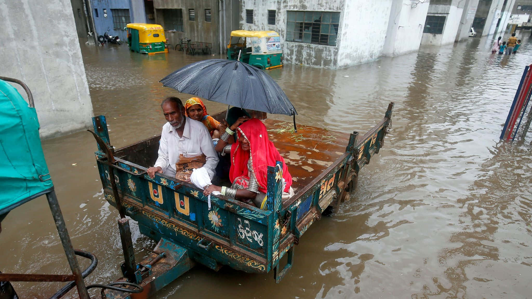 Flood 桌布 壁紙 — 三個人乘坐一輛裝飾過的卡車，在黑色雨傘下穿過泥濘的洪水，行駛在城市街道上。
