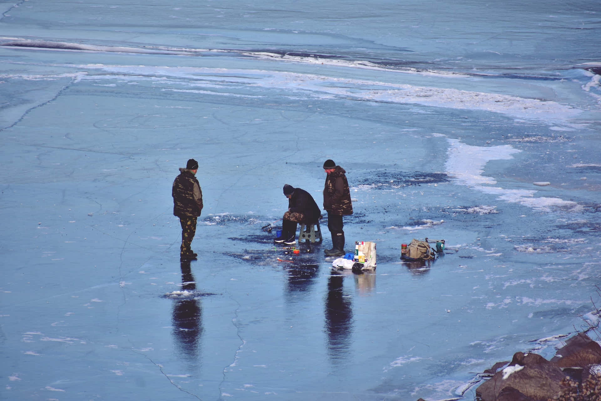Ice Fishing 桌布 壁紙 — 三個人在一個廣闊、裂開的藍色冰湖上冰釣，湖邊有灑滿雪的岩石。