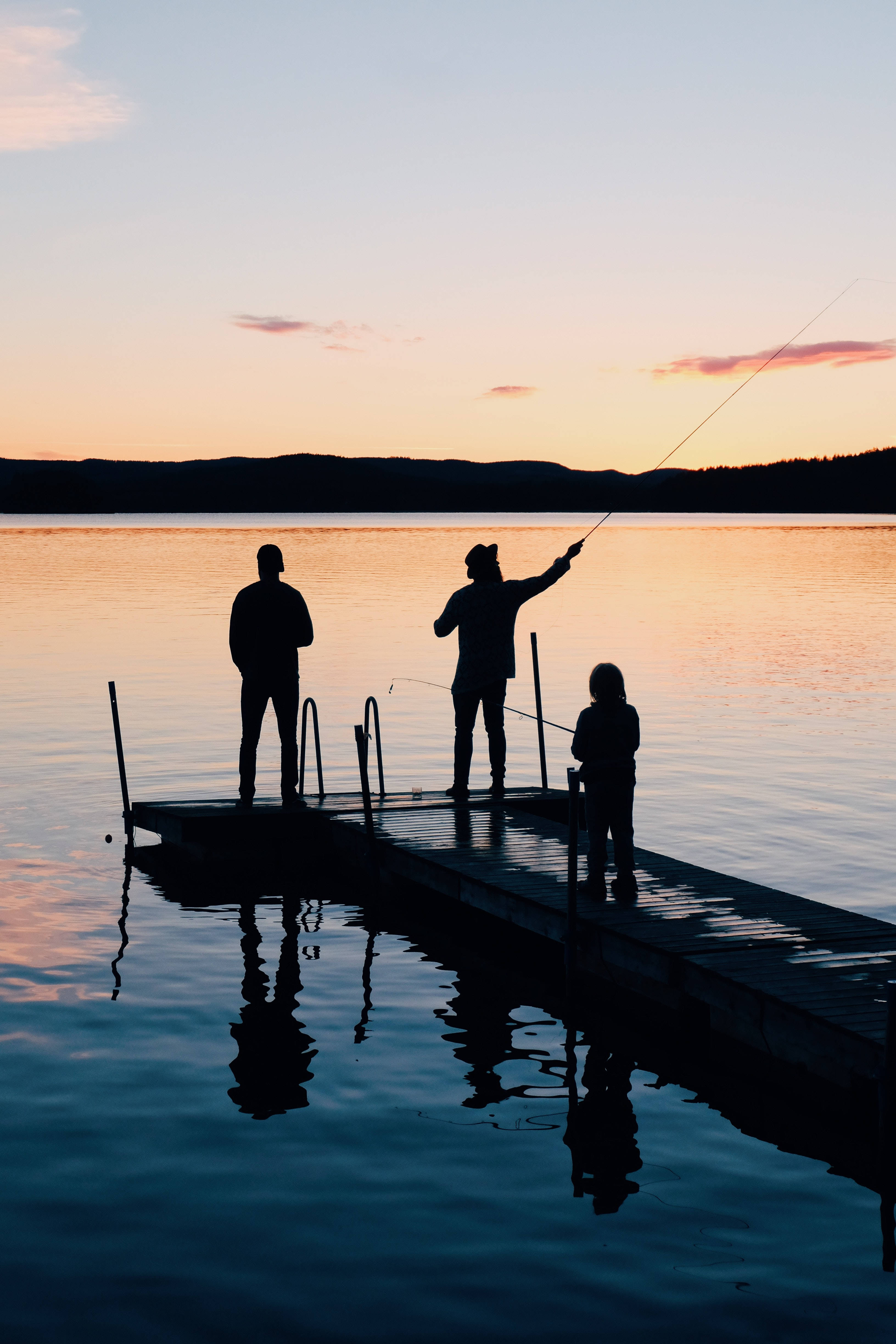 Family 桌布 壁紙 — 三個人在木碼頭上釣魚的剪影，夕陽西下，平靜的水面倒映著橙色的天空。