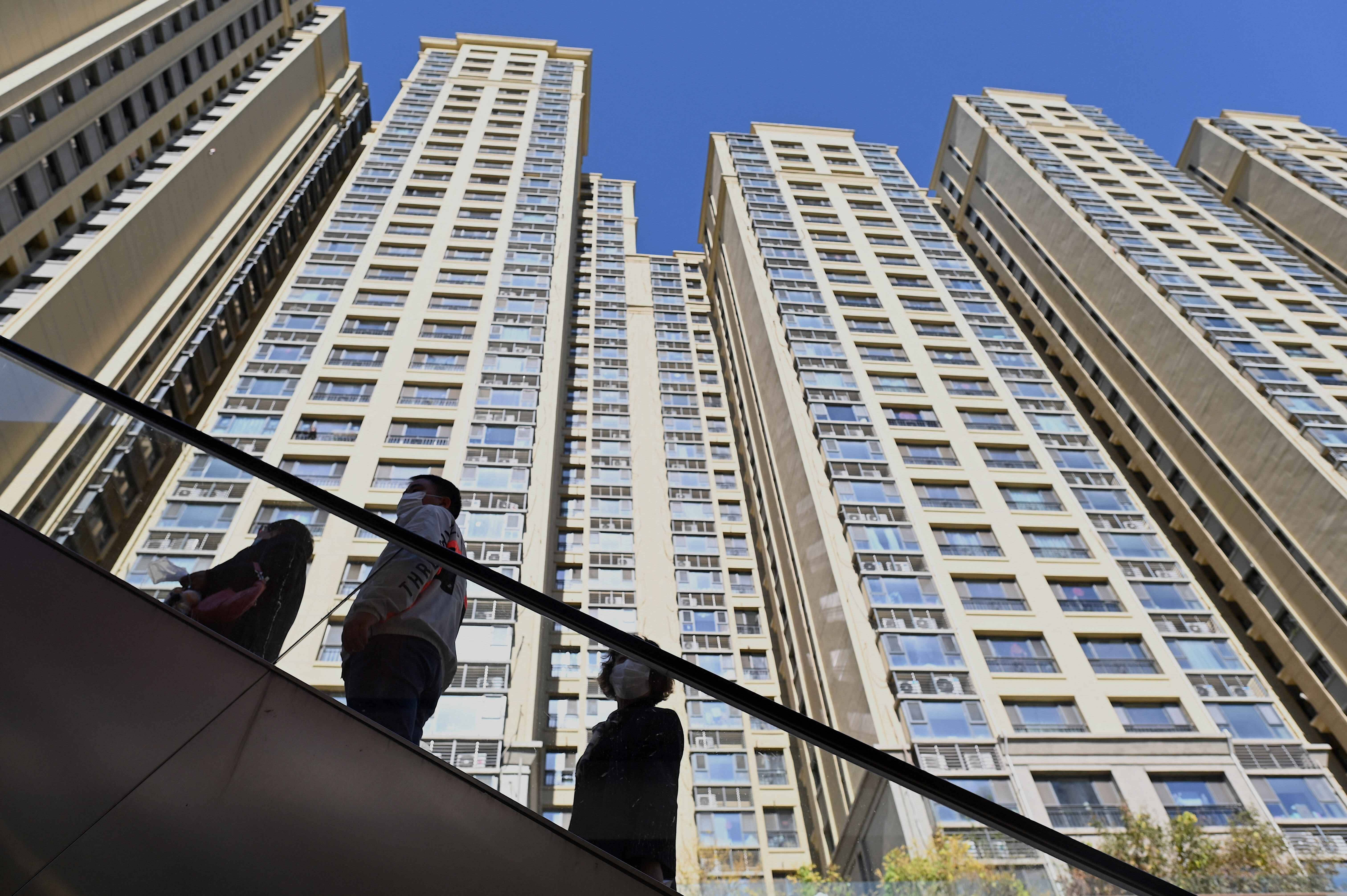 Buildings 桌布 壁紙 — 三個人搭乘手扶梯向上，在晴朗藍天下被高聳的米色公寓大樓襯托得渺小。