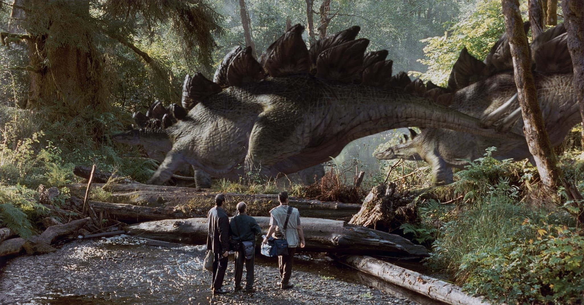 Jurassic Park Zoom 桌布 壁紙 — 三個人敬畏地站在森林溪床上，一隻巨大的劍龍在他們身後高聳的樹木中若隱若現。