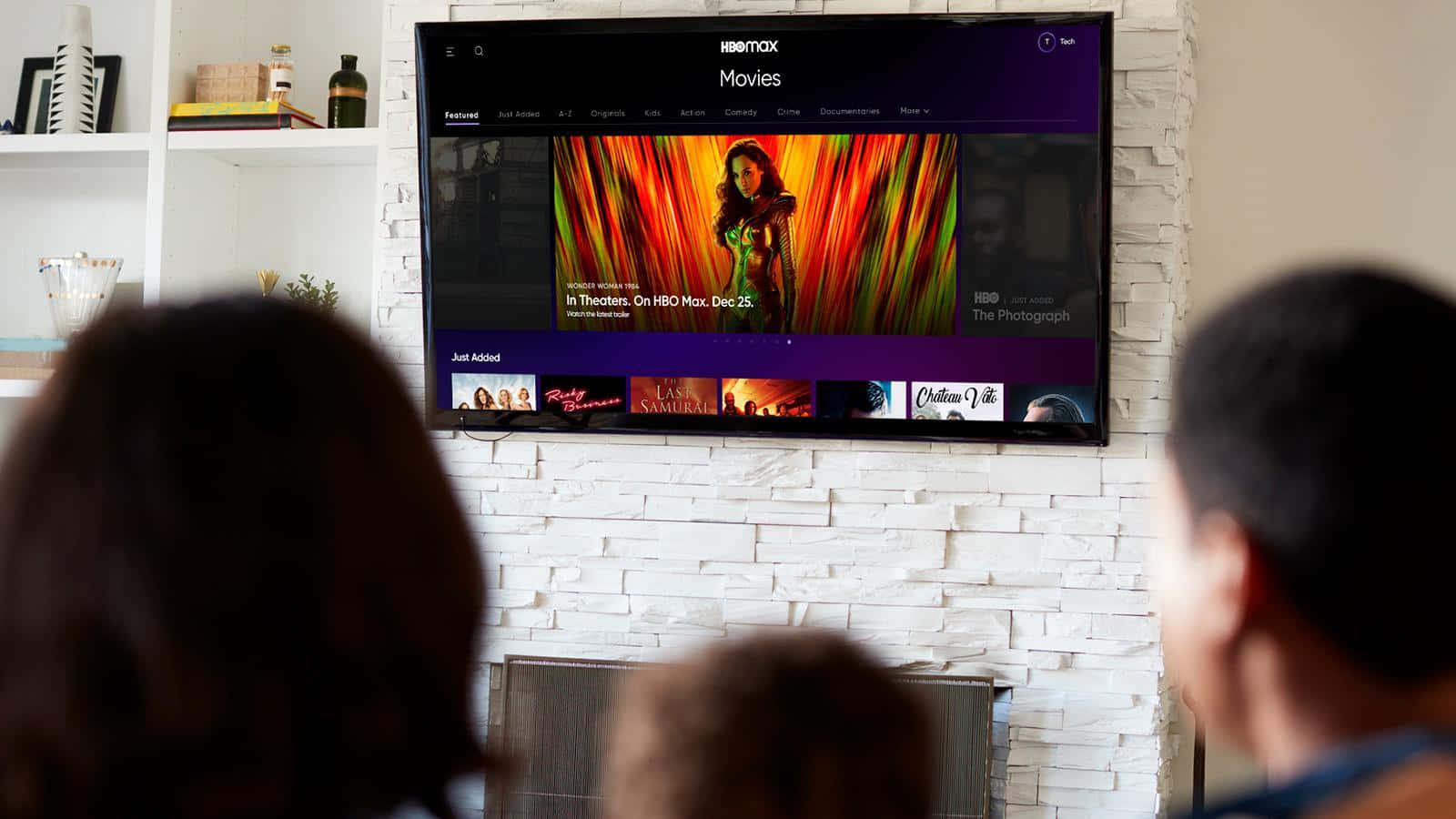 HBO 桌布 壁紙 — 三個人觀看安裝在白色磚砌壁爐上方的電視上的鮮豔HBO Max電影海報。