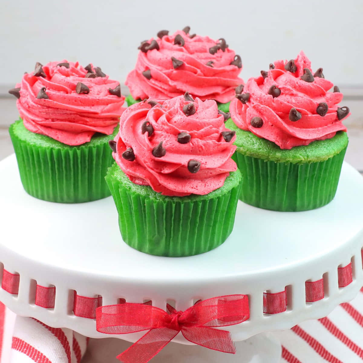 Pink Cupcakes 桌布 壁紙 — 三個以西瓜為主題的杯子蛋糕，綠色底座配上粉紅色糖霜，頂部綴有巧克力豆，放在一個飾有紅色絲帶的白色蛋糕架上。