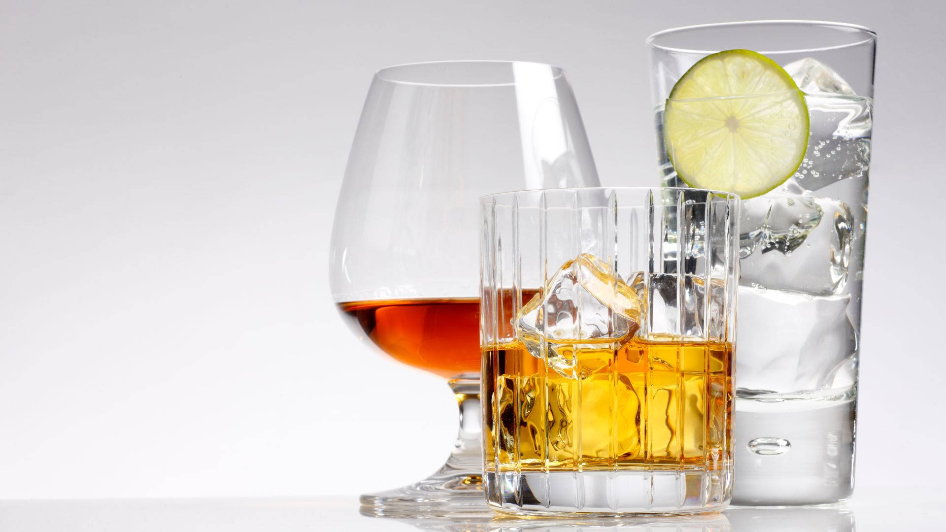 alcohol 桌布 壁紙 — 三個優雅的玻璃杯，裝滿琥珀色威士忌和清澈的雞尾酒，飾以萊姆片和冰塊，置於潔白的表面上。