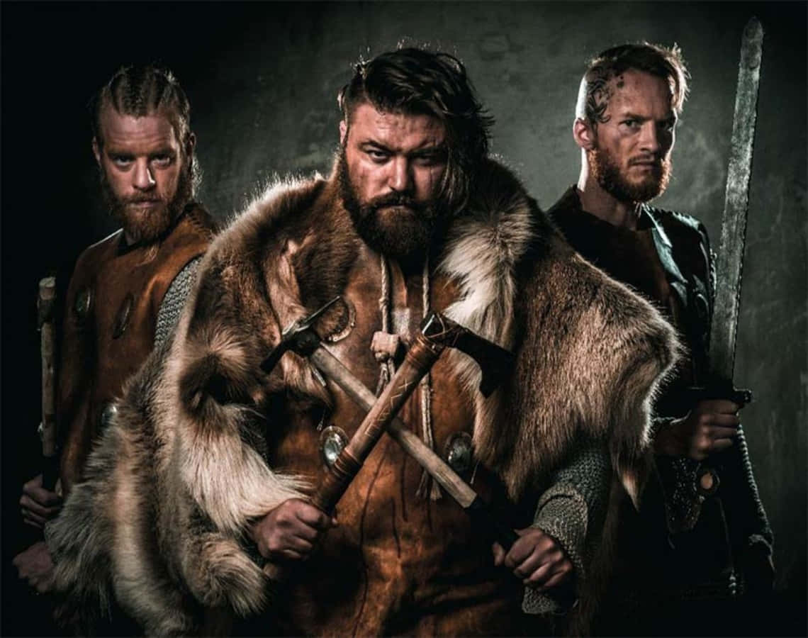 Vikings 桌布 壁紙 — 三個兇猛的維京人身穿毛皮斗篷和盔甲，手持斧頭和劍，在黑暗粗糙的背景中威嚇地站立。