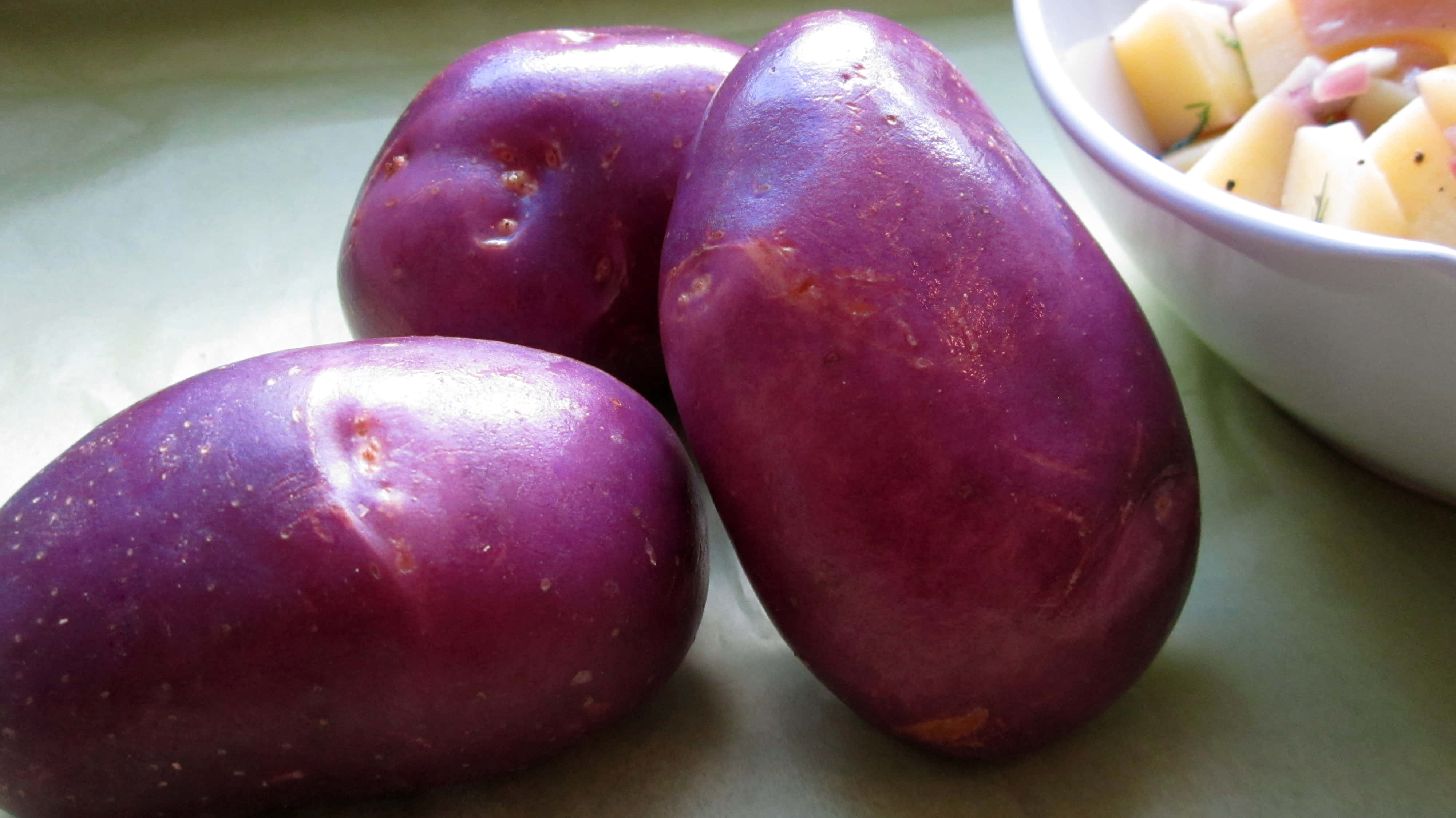 Purple Potato 桌布 壁紙 — 三個光澤的紫色馬鈴薯放在淺綠色表面上，旁邊有一碗馬鈴薯丁沙拉。
