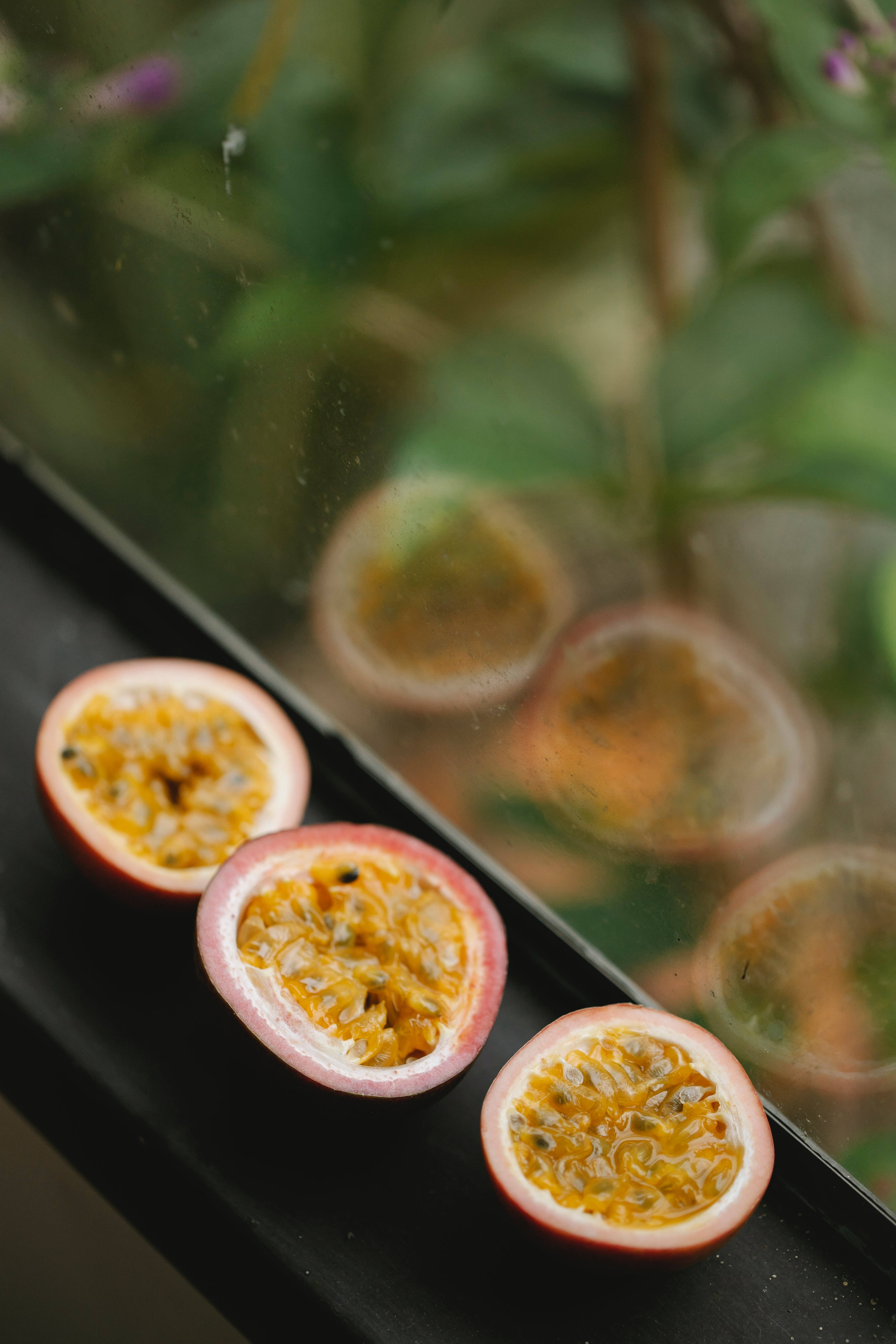 Fruits 桌布 壁紙 — 三個切半的百香果，帶有鮮黃色的果肉和黑色的種子，放在一個黑暗的表面上，旁邊是一個反射模糊綠意的玻璃杯。