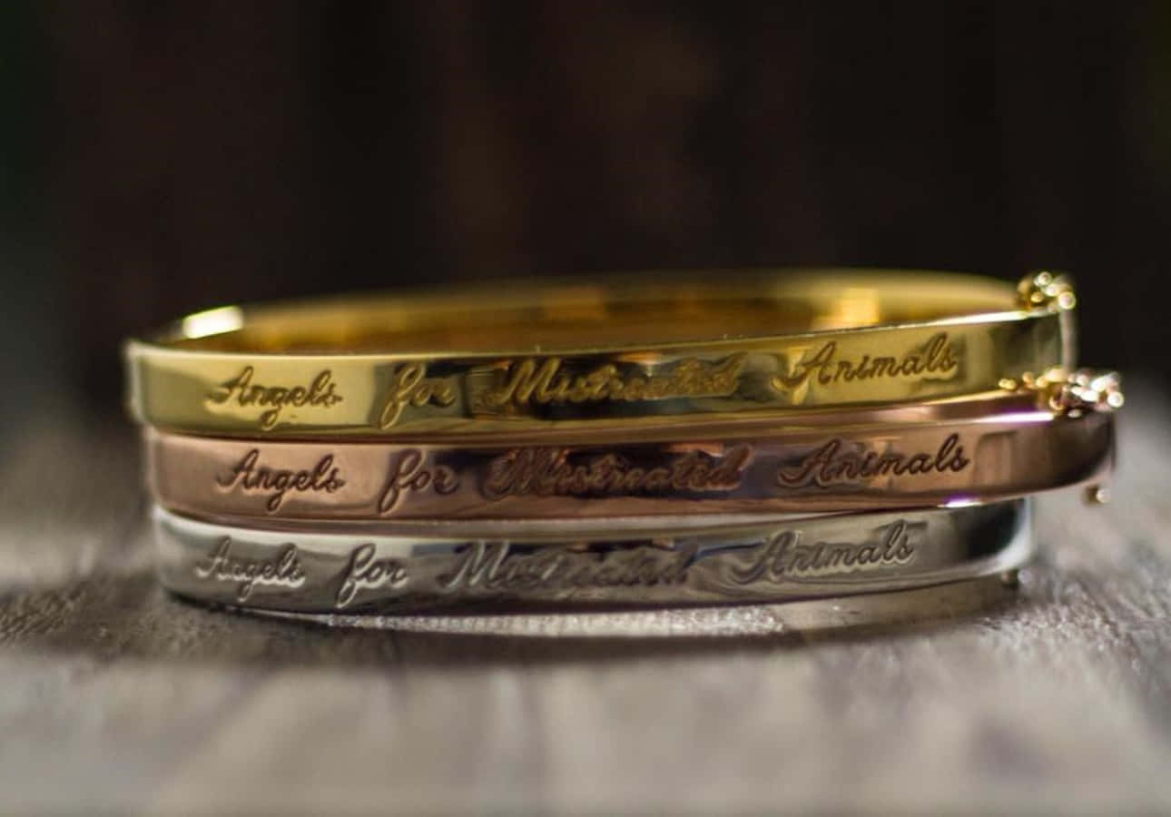 Bangles 桌布 壁紙 — 三個刻有「Angels for Mistreated Animals」的金屬手鐲疊放在有紋理的表面上，光線柔和。