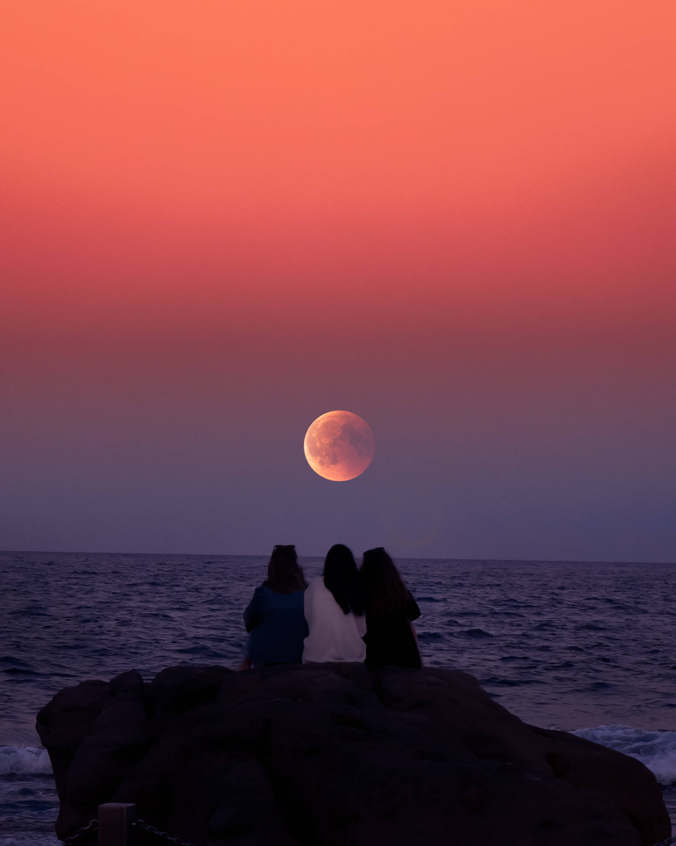full moon 桌布 壁紙 — 三個剪影人物坐在海邊岩石上，觀賞黃昏時一輪發光的橙色月亮從水面升起。