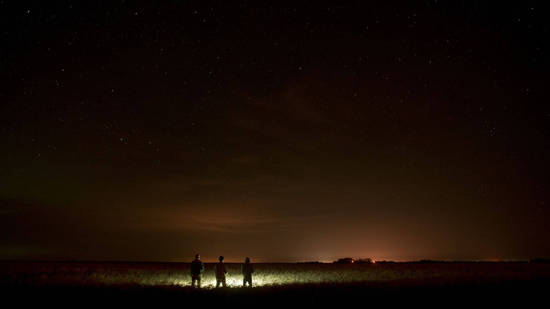 Best Dark 桌布 壁紙 — 三個剪影人物站在草地上，頭上是廣闊的星空，地平線遠處有橙色的光芒。
