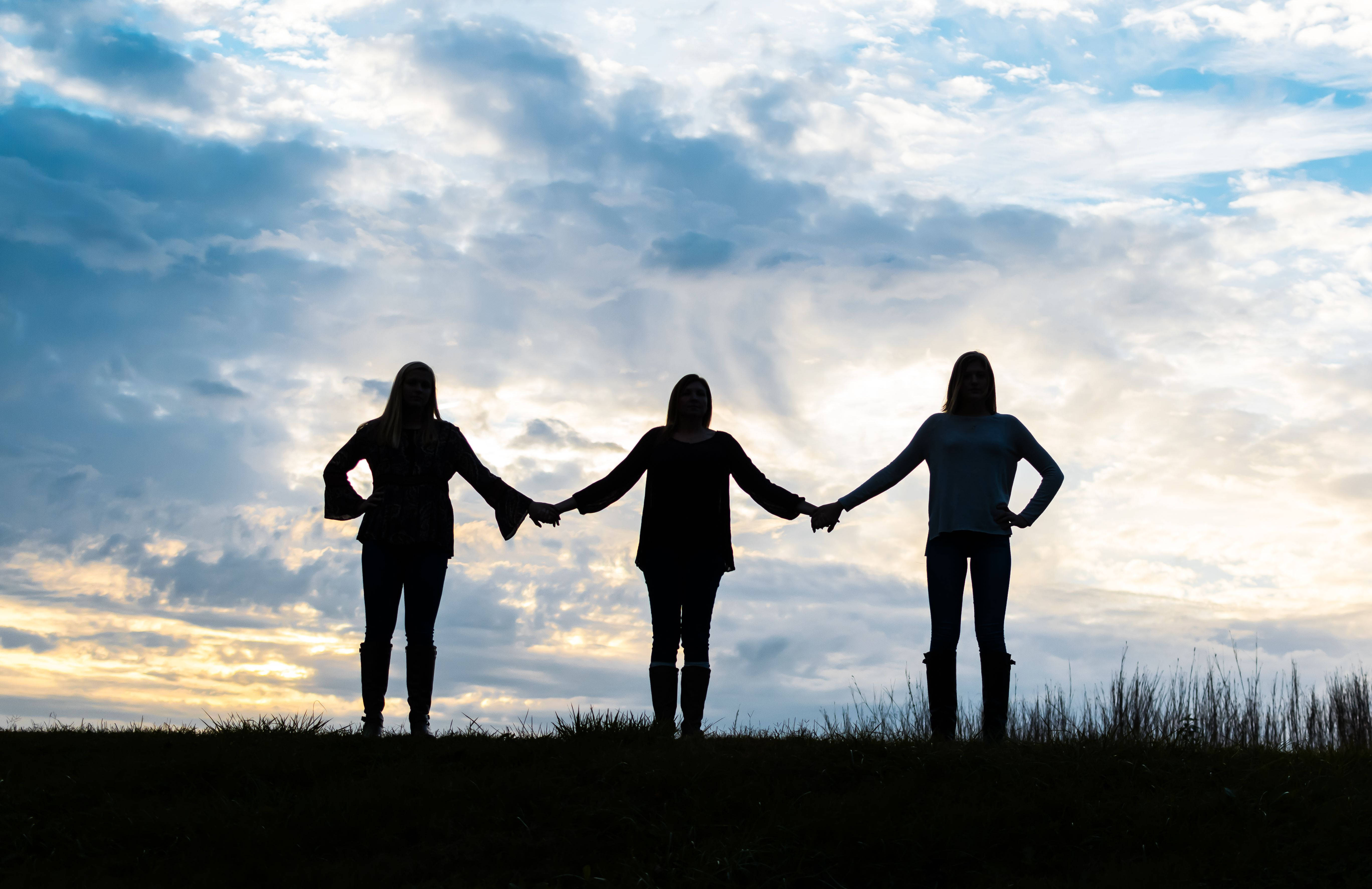 3 Best Friends 桌布 壁紙 — 三個剪影女性在長滿草的山丘頂上排成一線手牽手，背景是戲劇性的黃昏天空。