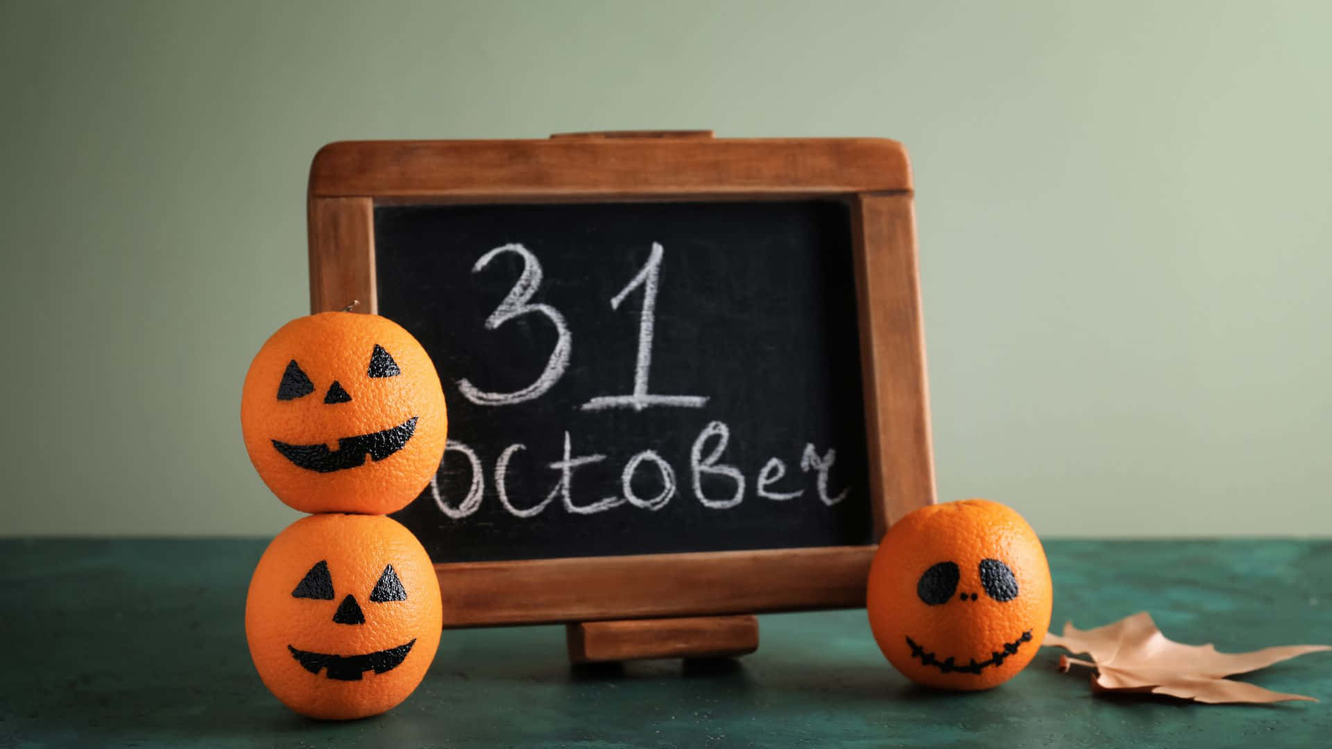 October 31st 桌布 壁紙 — 三個南瓜燈橙子放在一塊顯示10月31日的黑板前，背景為柔和的綠色。