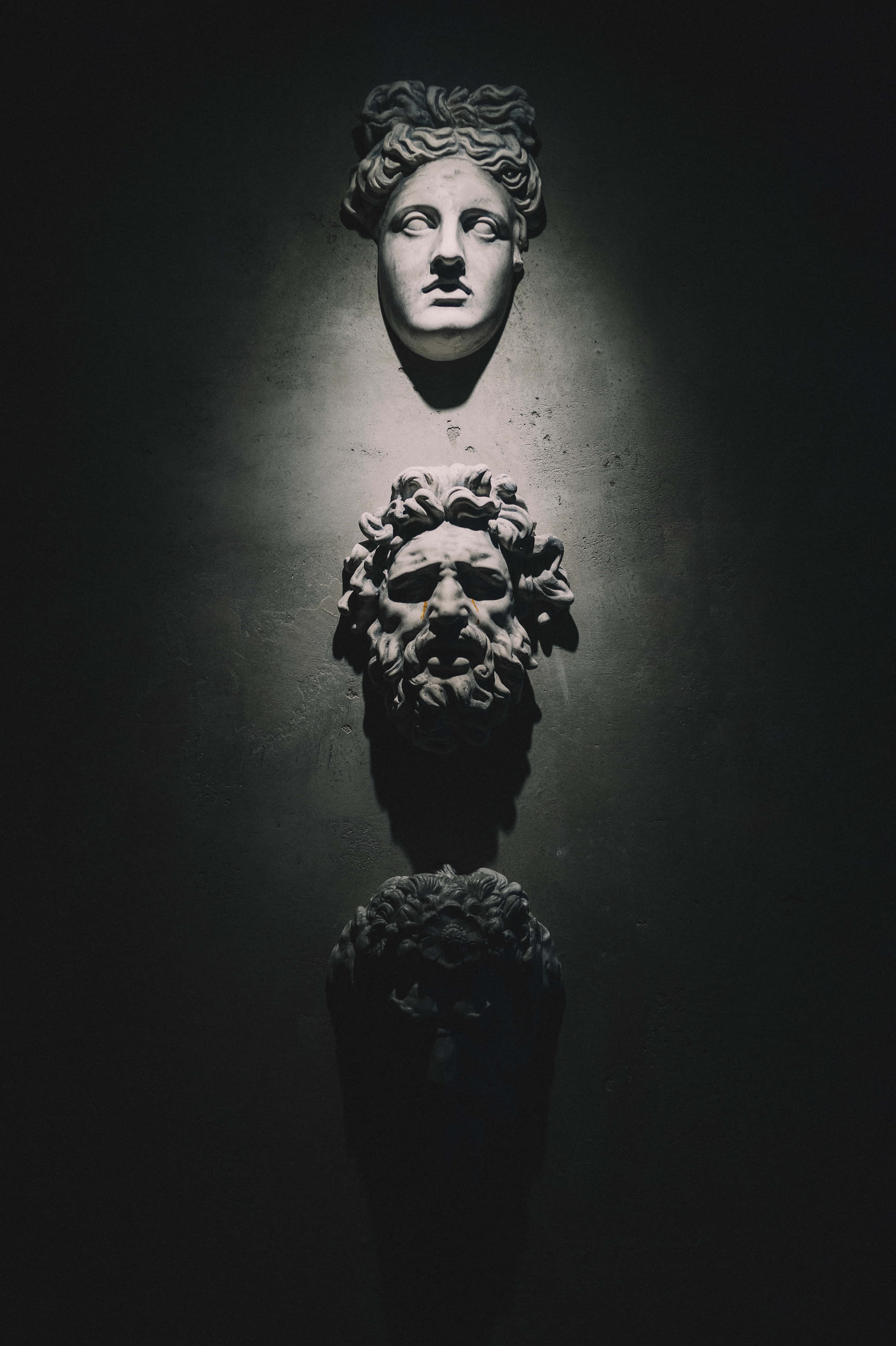 Best Greek Mythology 桌布 壁紙 — 三個古典石像半身像垂直懸掛在昏暗的房間中，由單一聚光燈照亮，投射出戲劇性的陰影。