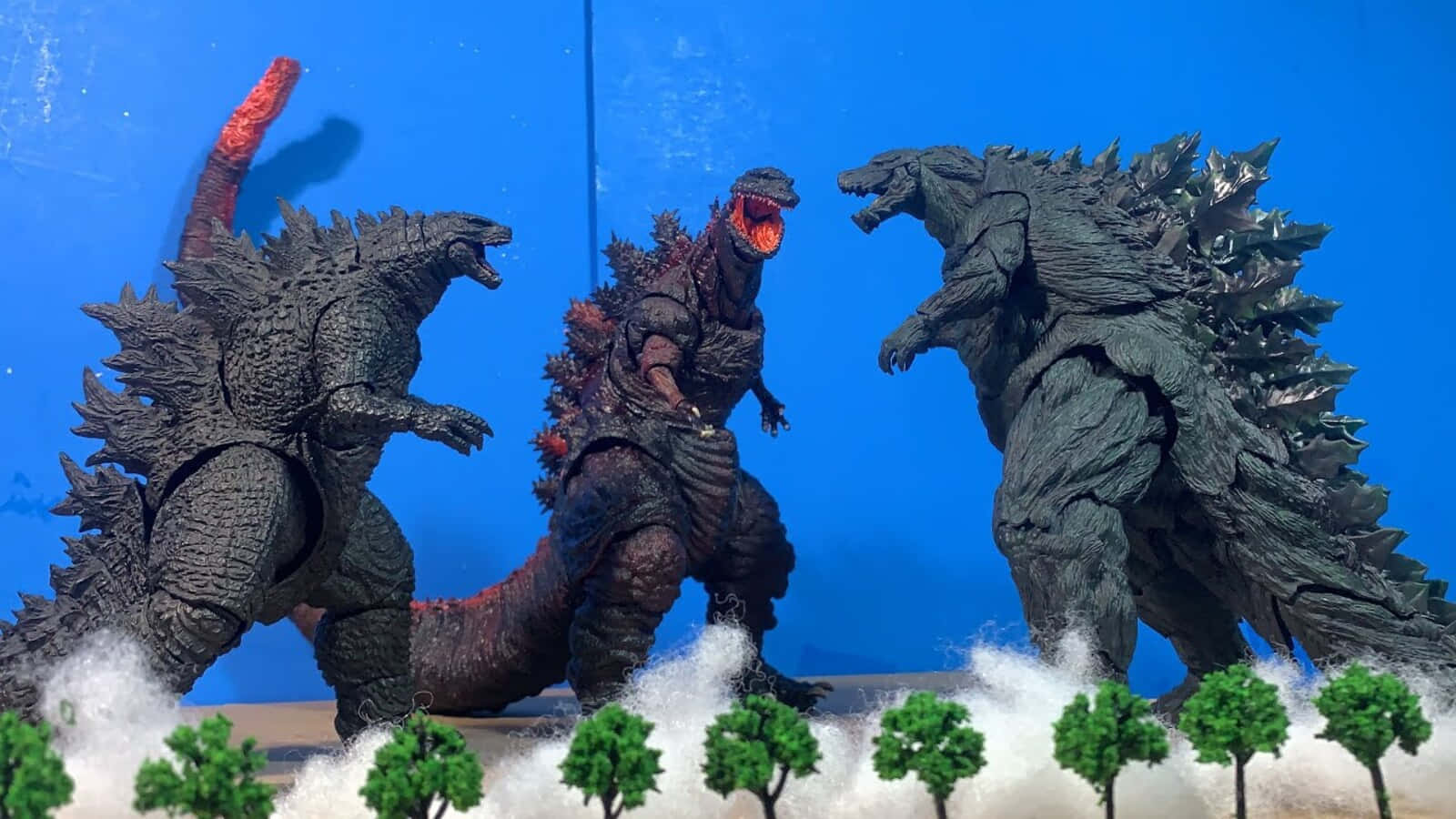 Godzilla Earth 桌布 壁紙 — 三個哥吉拉動作人偶以戰鬥姿勢站立在雪地地形上，配有微型樹木，背景為純藍色。
