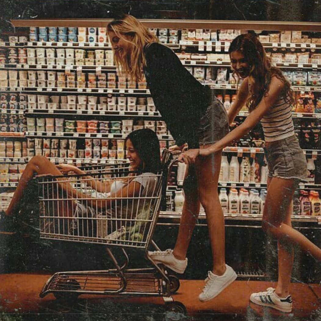 Friends 桌布 壁紙 — 三個大笑的朋友在雜貨店裡嬉戲互動，一個女孩坐在購物車裡，由另外兩個人推著。