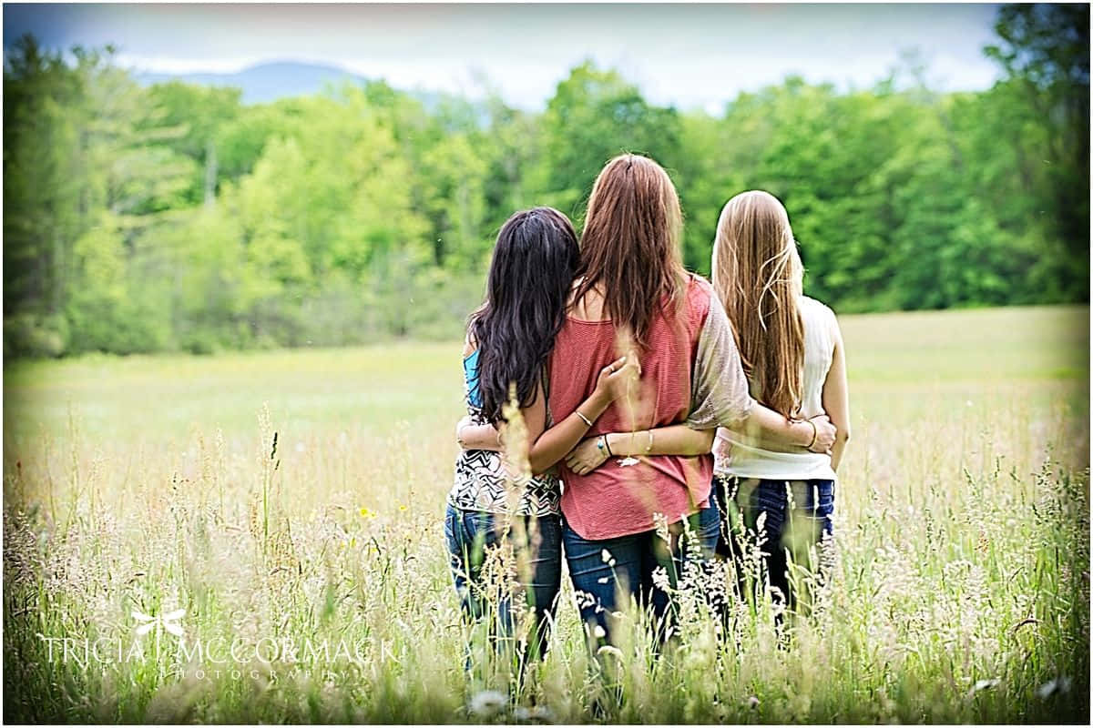 Friends 桌布 壁紙 — 三個女孩在陽光明媚的高草田野中擁抱，背景是樹木繁茂的山丘。