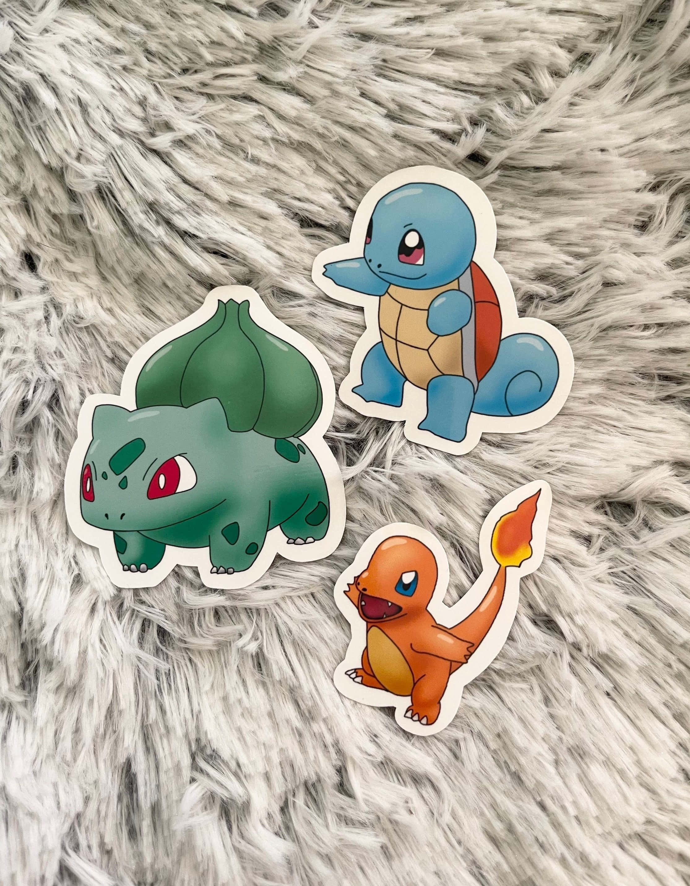 Pokemon Stickers 桌布 壁紙 — 三個妙蛙種子、傑尼龜和小火龍的寶可夢貼紙放在一張毛茸茸的灰色地毯上。