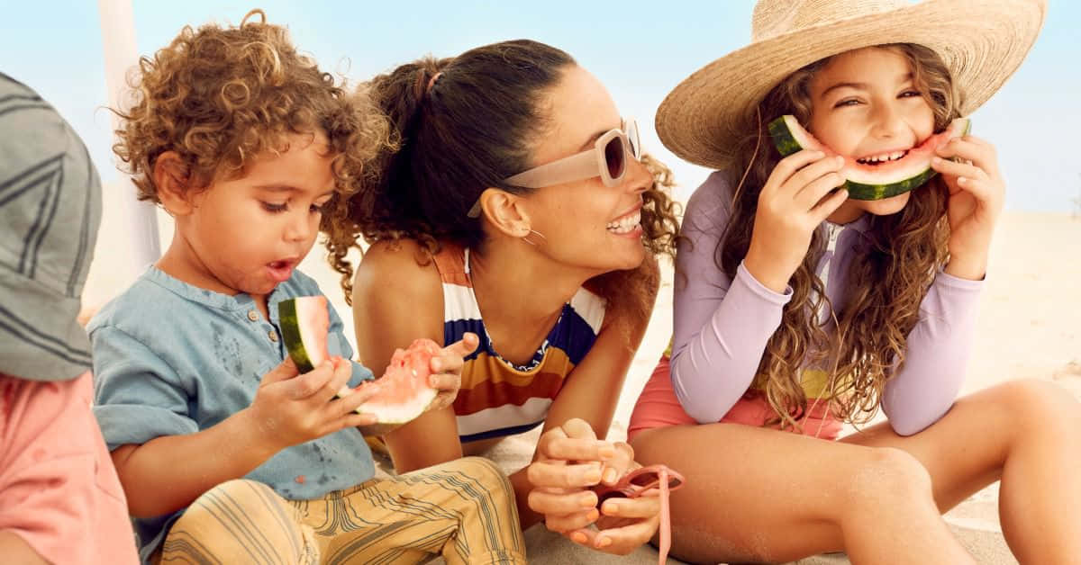 Beach Family 桌布 壁紙 — 三個孩子和一個女人在陽光明媚的海灘上笑著吃西瓜片。