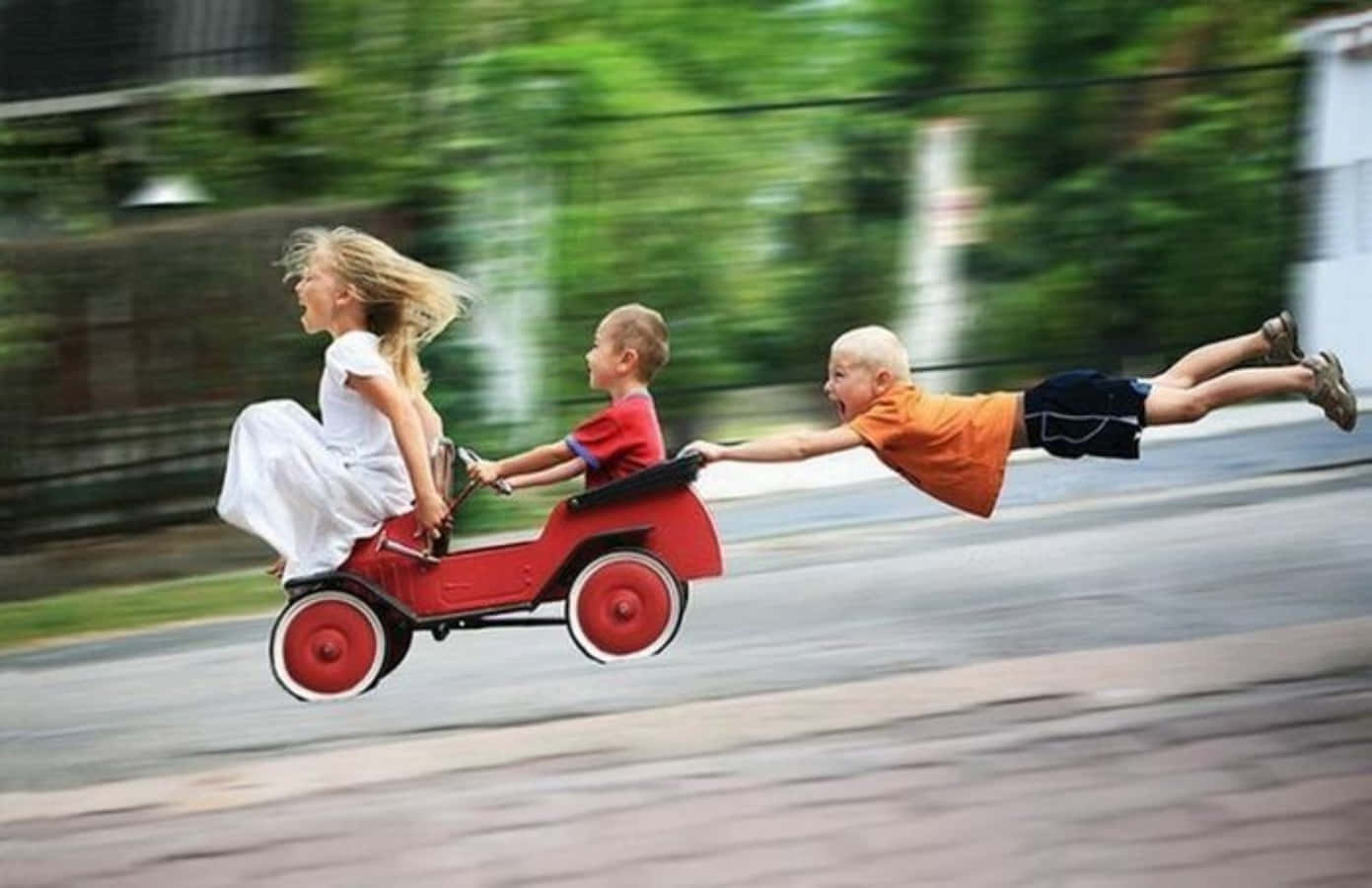 小孩動圖 桌布 壁紙 — 三個孩子在模糊的郊區街道上比賽紅色玩具車，一個男孩在後面飛翔。
