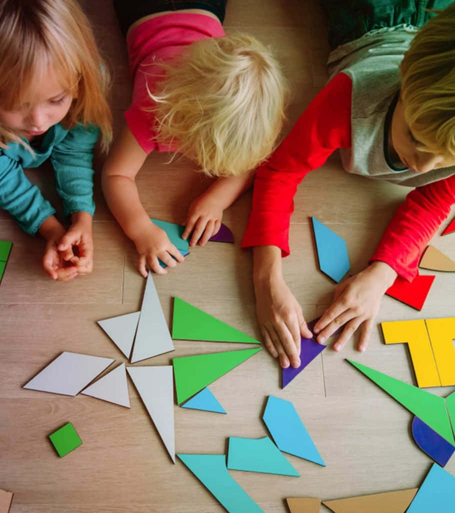數學 桌布 壁紙 — 三個孩子躺在木地板上，專注地將色彩繽紛的幾何拼圖塊排列在他們周圍。