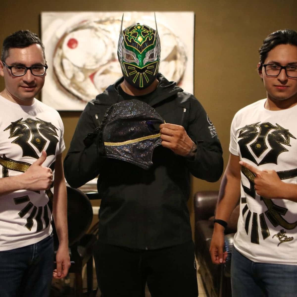 WWE 桌布 壁紙 — 三個室內男子，一人戴著綠色金色 lucha libre 面具，手持另一個面具，另外兩人穿著相配的龍 T 恤豎起大拇指。